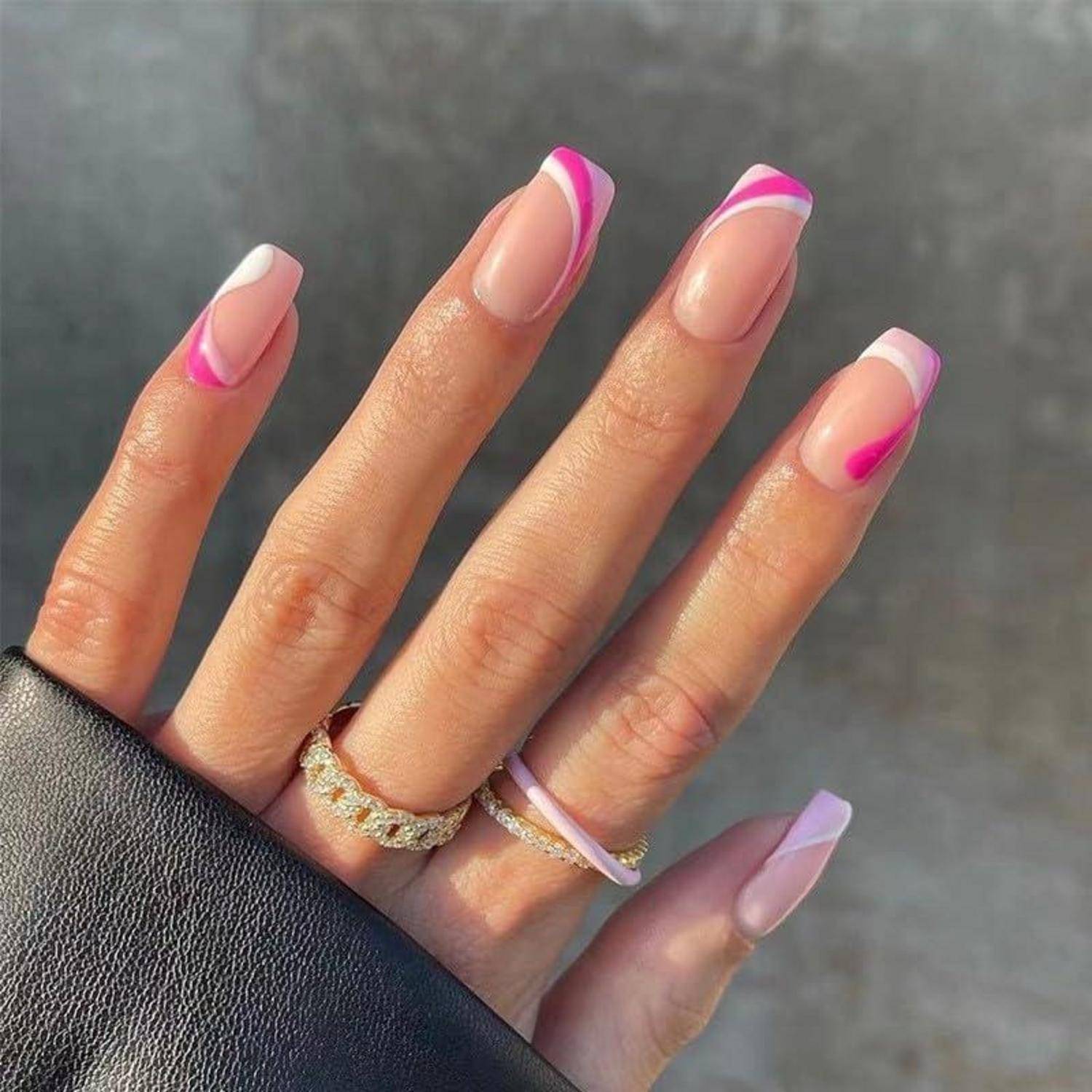 Hand mit langen mandelförmigen Nägeln in Nude-Lack und rosa-weißen Spitzen, mit Gold- und Lavendel-Ringen vor grauem Hintergrund.