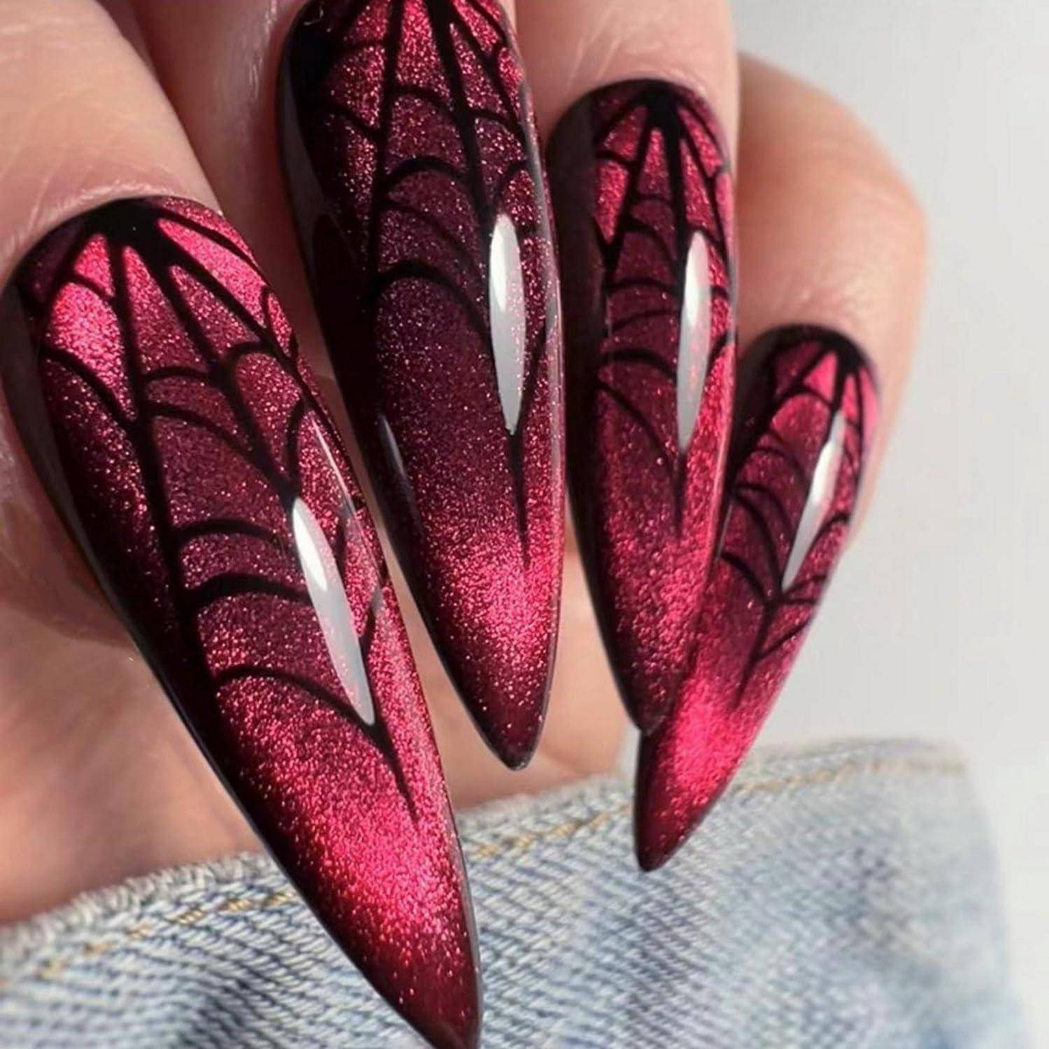 Nahaufnahme von langen Stiletto-Nägeln mit glänzend rotem Glitter und schwarzem Spinnennetz-Nageldesign auf einer durchscheinenden rosa Basis.