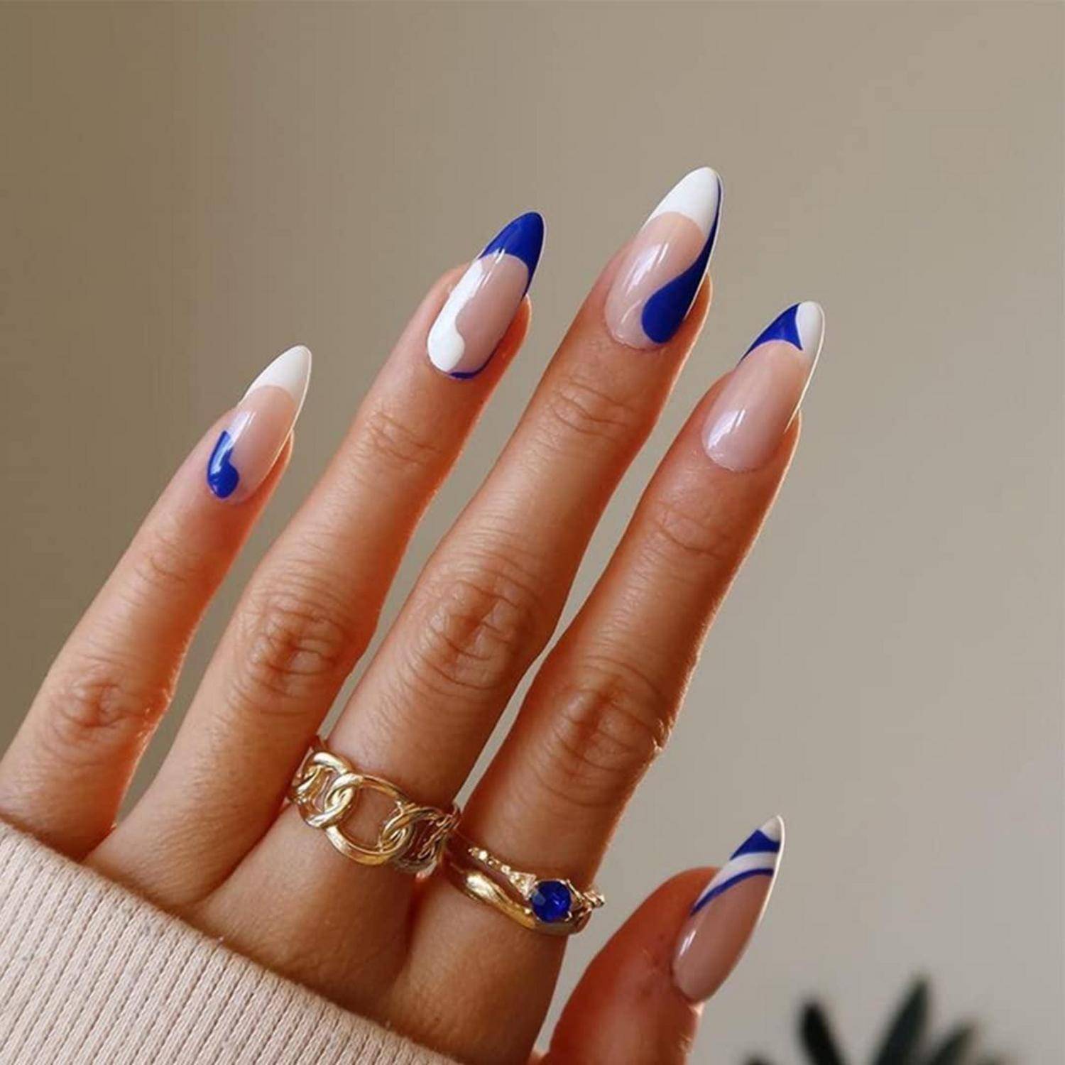 Hand mit langen mandelförmigen Nägeln in Nude, Weiß und Blau im French-Tip-Design, mit goldenen Ringen vor einem einfarbigen Hintergrund.