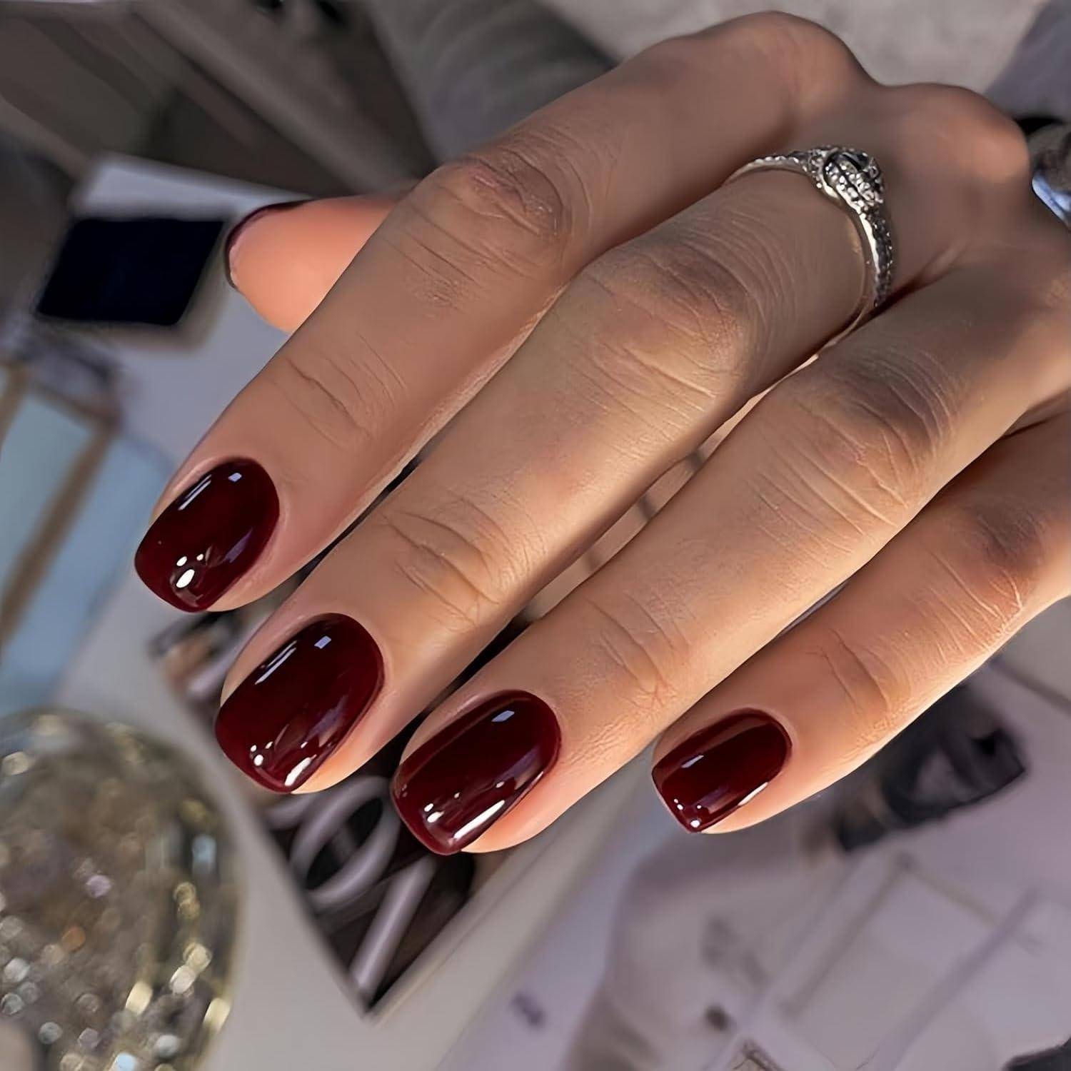 Nahaufnahme einer Hand mit kurzem, glänzendem dunkelrot-burgunderfarbenem Nageldesign und einem silbernen Ring.