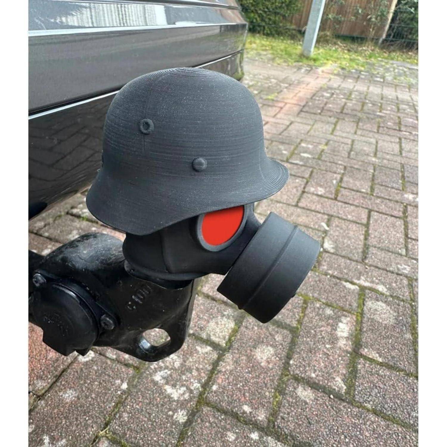 Anhängerkupplung mit einem schwarzen Novelty-Helm und Gasmaske, daneben ein geparktes Fahrzeug auf einer Ziegsteineinfahrt.