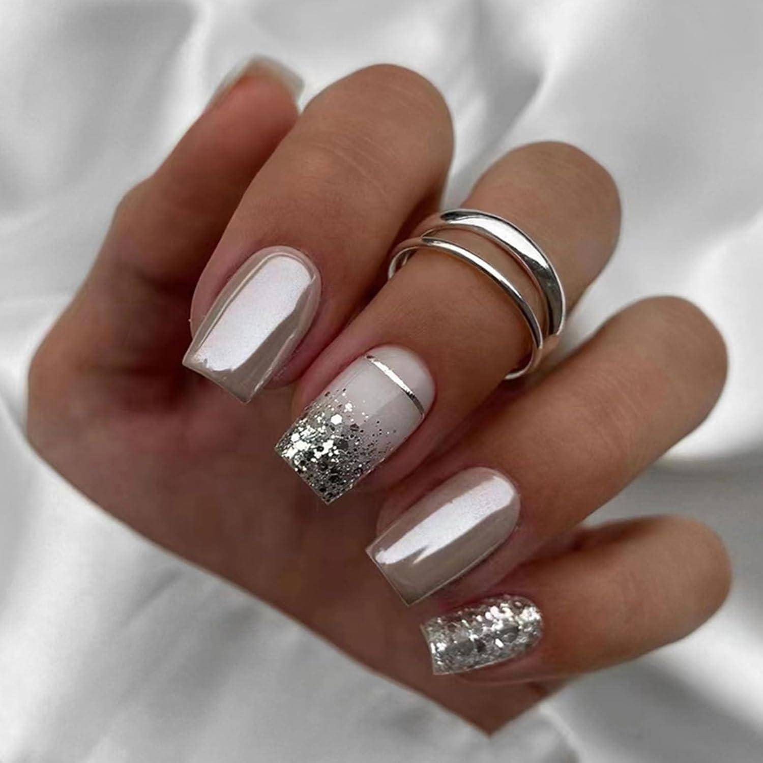 Nahaufnahme einer Hand mit glänzender, nackter, quadratischer Maniküre mit silbernem Glitter-Akzentnägeln und dünnen silbernen Ringen.