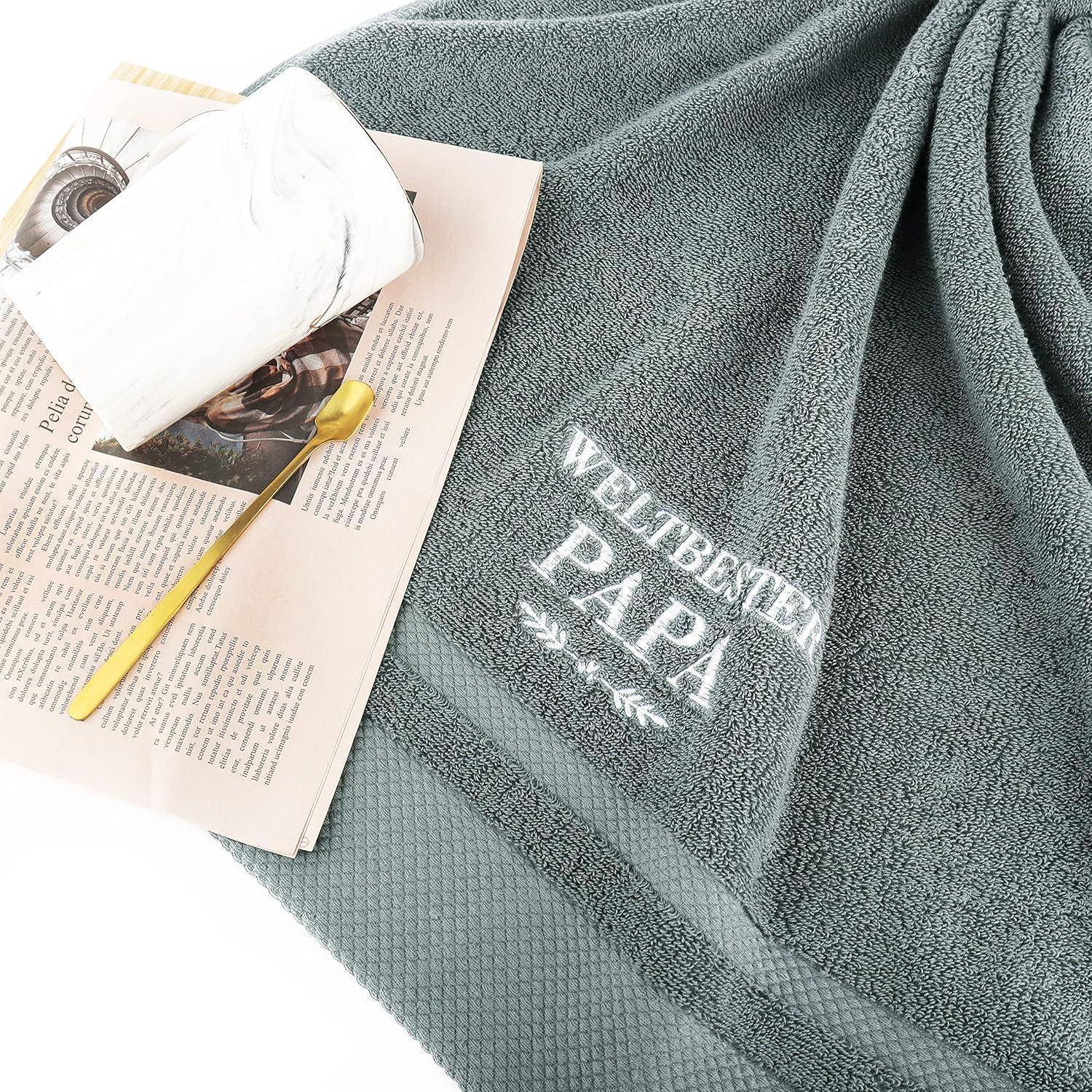 Graue strukturierte Decke mit besticktem 'WELTBESTE PAPA' neben einem aufgeschlagenen Magazin, weißer Tasse und gelbem Löffel auf einer weißen Oberfläche.