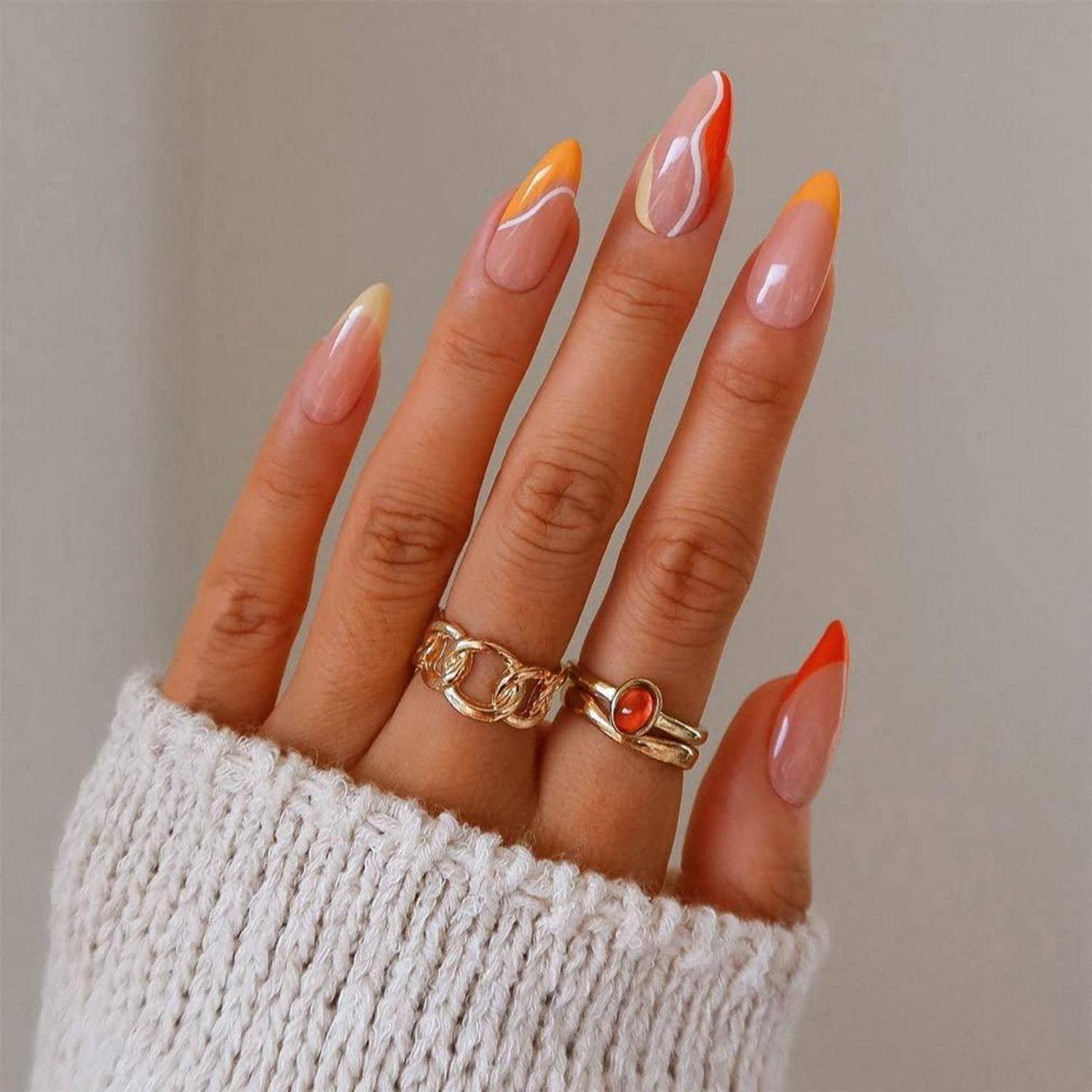 Nahaufnahme einer Hand mit langen mandelförmigen Nägeln in Rosa und Orange mit französischen Spitzen, goldenen Ringen und Manschette eines weißen Strickpullovers.