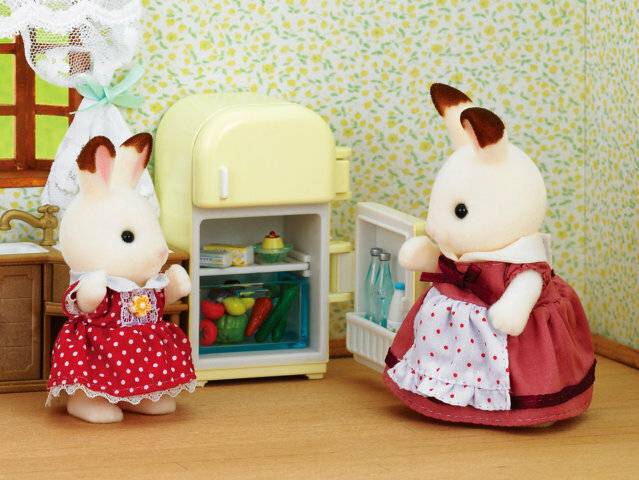 Zwei Kaninchen-Figuren in einer Spielküche, eine hält einen Apfel in der Nähe eines offenen Kühlschranks, während die andere daneben in einer Schürze steht.