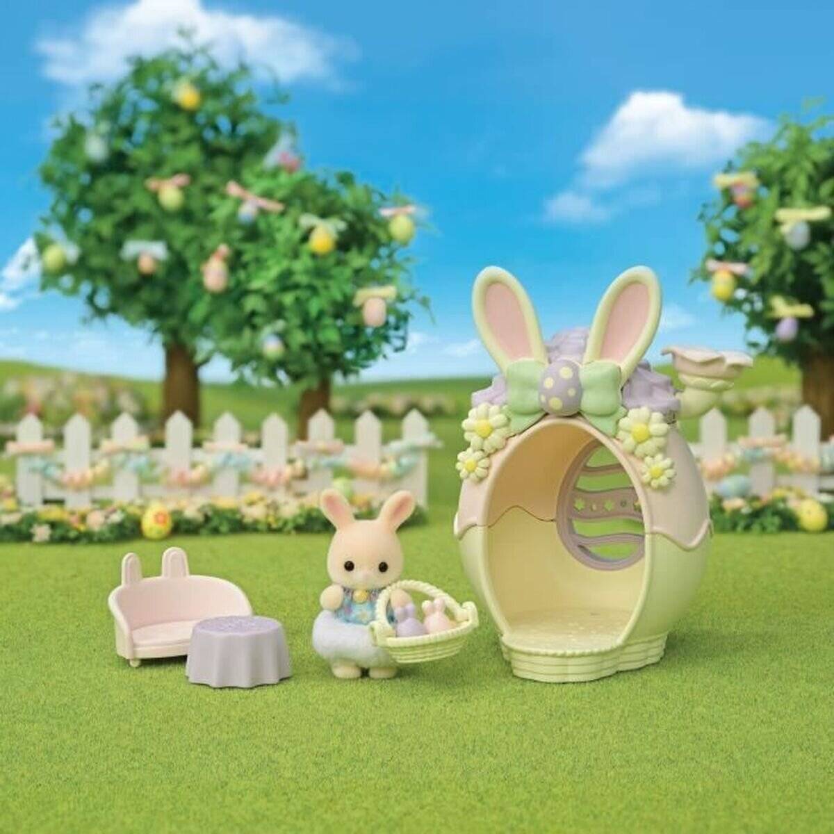 Spielzeughase mit einem Osterkorb steht neben einem großen eiförmigen Haus und einem kleinen Tisch- und Stuhtsatz in einer sonnigen Gartenszene.