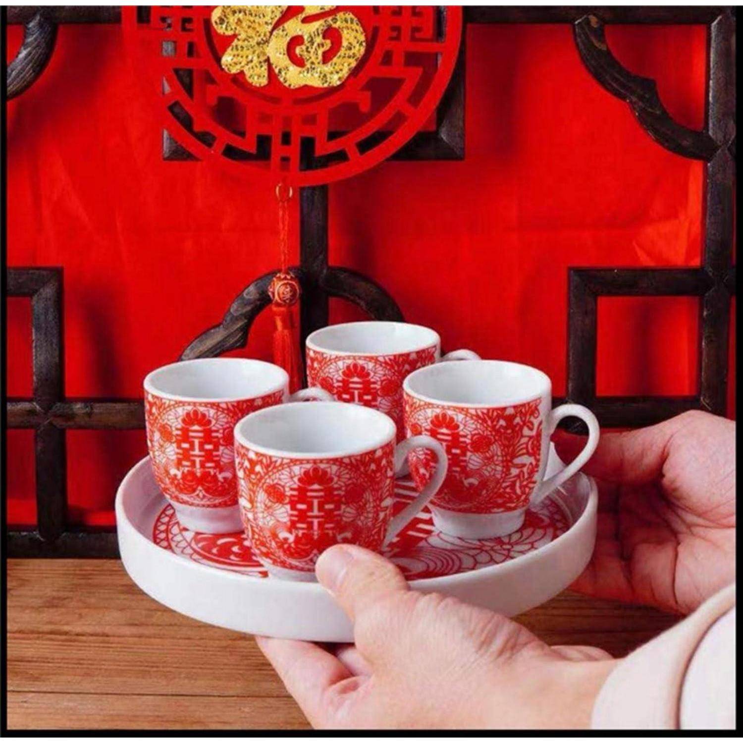 Hände präsentieren ein weißes Tablett mit vier rot-weißen chinesischen Teetassen und Untertassen, gezeigt vor einem festlichen roten Hintergrund mit einem dekorativen Symbol.
