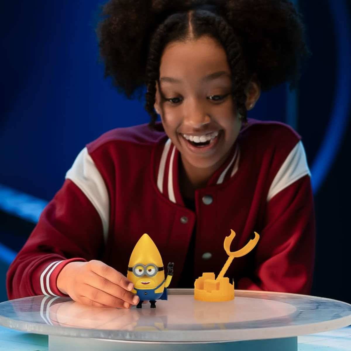 Kind mit einem Lächeln in einer roten Universitätsjacke, das eine kleine Minion-Figur neben einem gelben krallenförmigen Spielzeug auf einem transparenten runden Tisch zurechtrückt.
