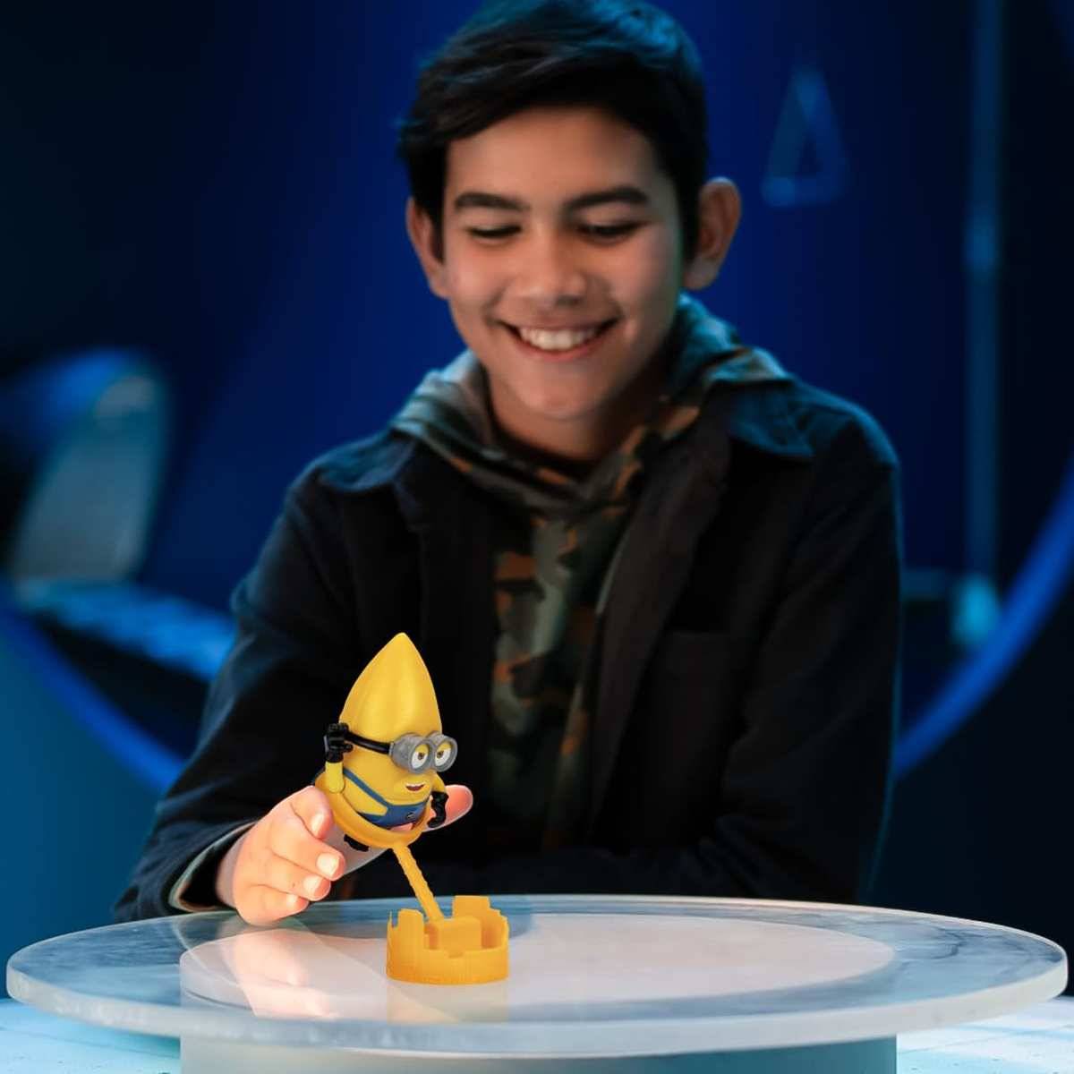 Ein lächelnder Junge spielt mit einer Minion-Figur auf einer Tischplatte und setzt sie in einen kleinen gelben Sockel in einem blau beleuchteten Raum.