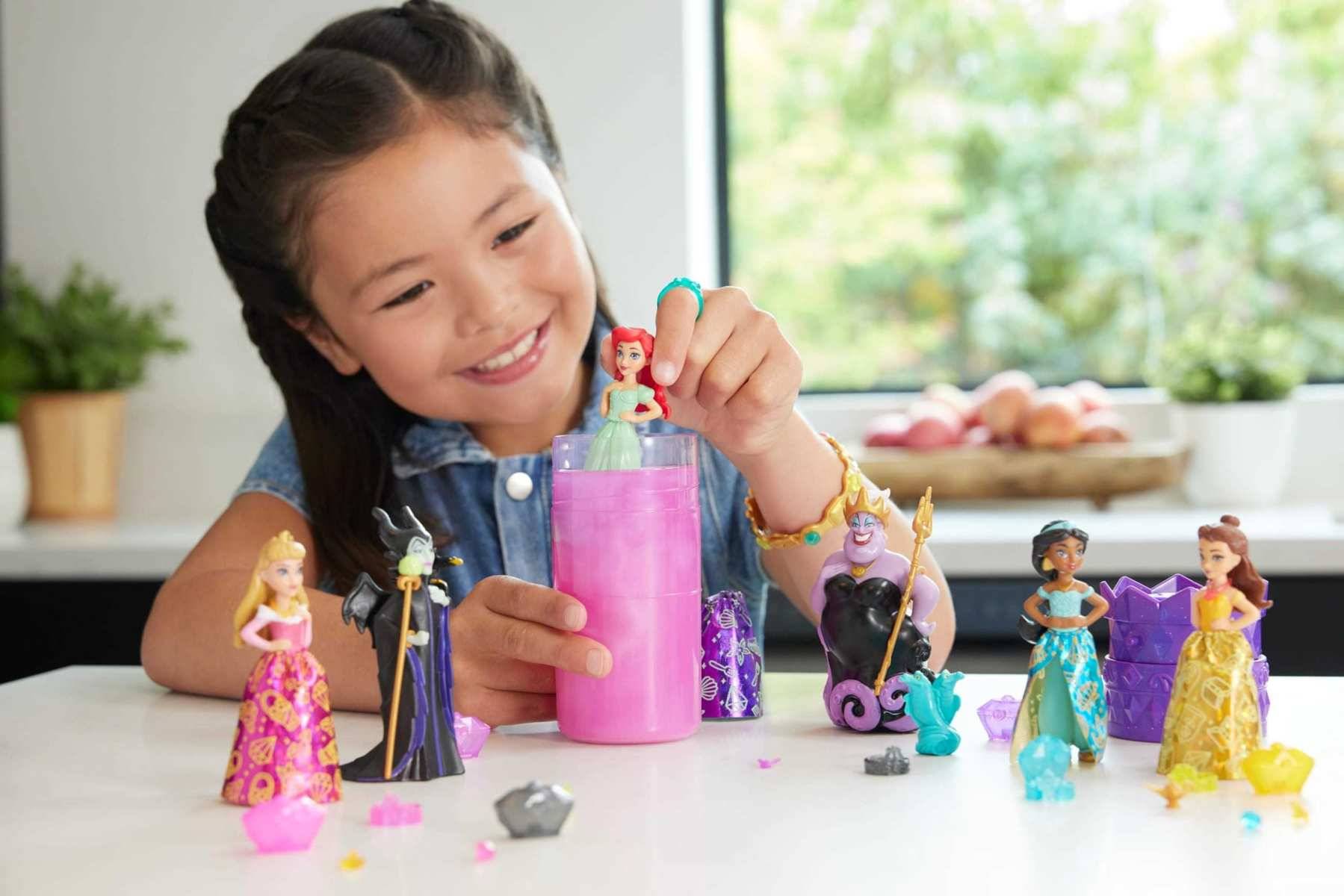 Ein lächelndes Mädchen spielt mit einem rosa Spielzeugbehälter und mehreren kleinen Prinzessinnenfiguren auf einem Tisch im Innenbereich.