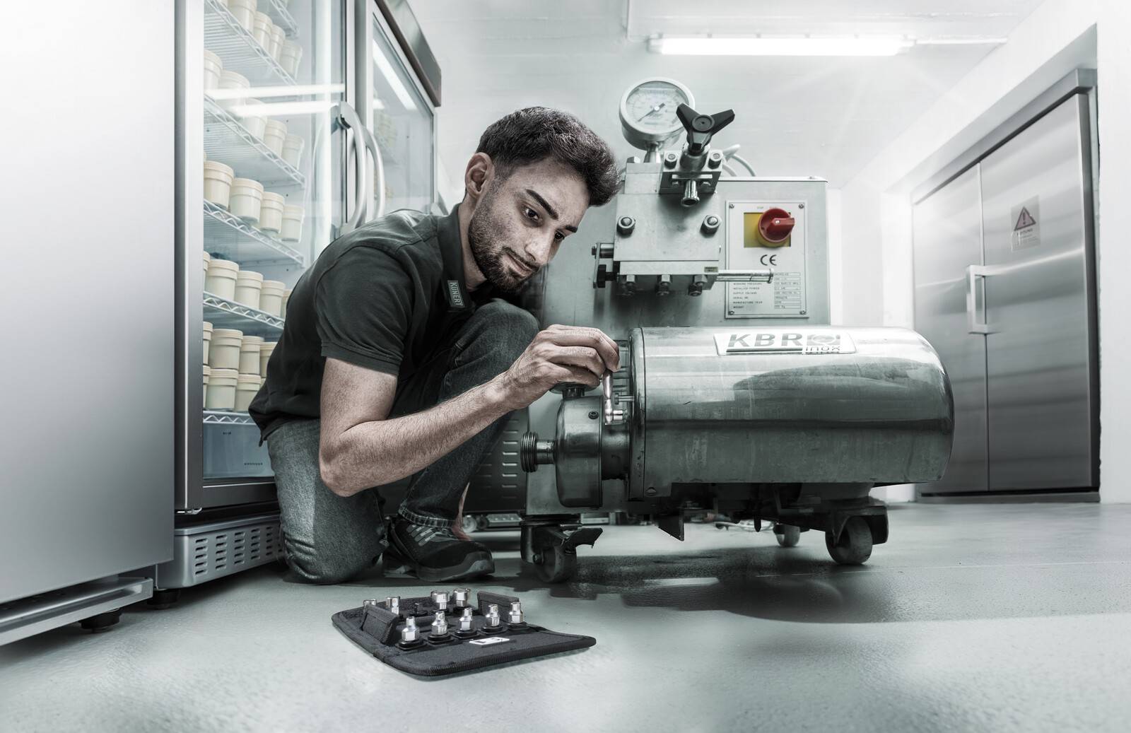 Ein Mann kniet neben einer Edelstahl-Industriemaschine in einem sauberen Raum und stellt eine Seitenbuchse in der Nähe eines Bedienfelds ein.