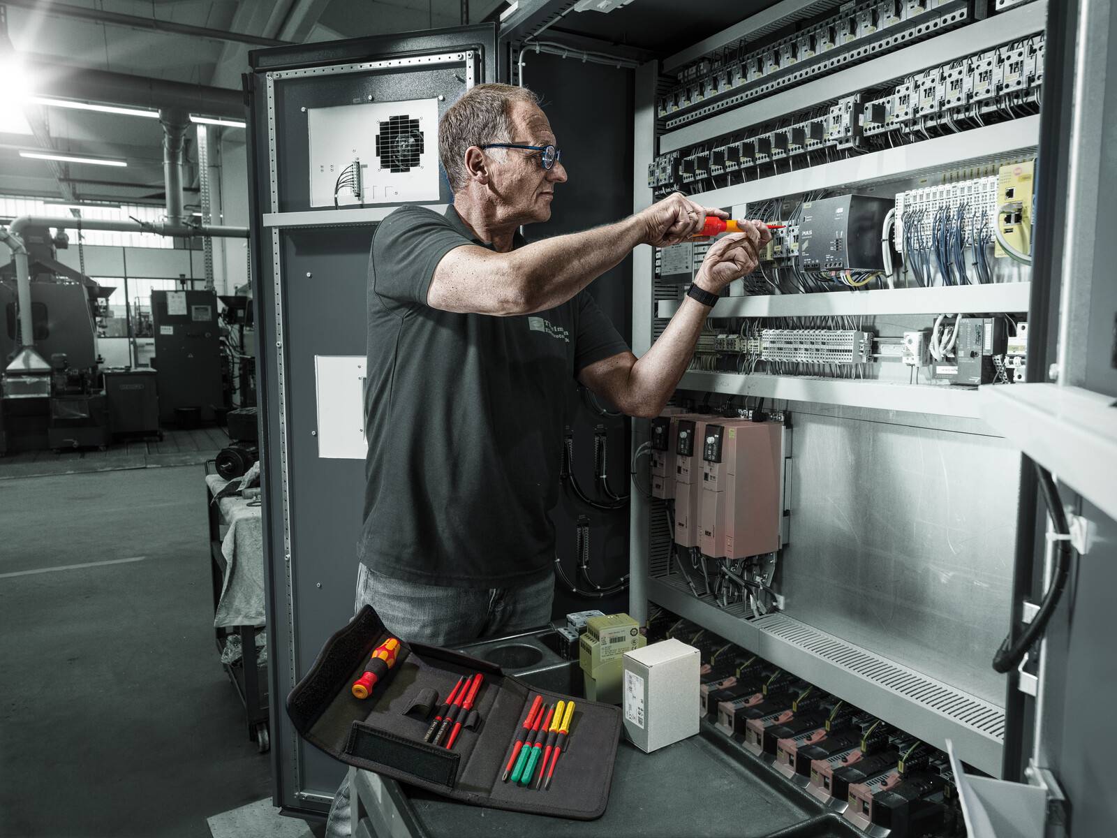 Techniker, der mit einem Schraubendreher einen offenen elektrischen Schaltschrank in einer Industrieanlage wartet, mit Werkzeugen, die auf einem Tablett ausgelegt sind.