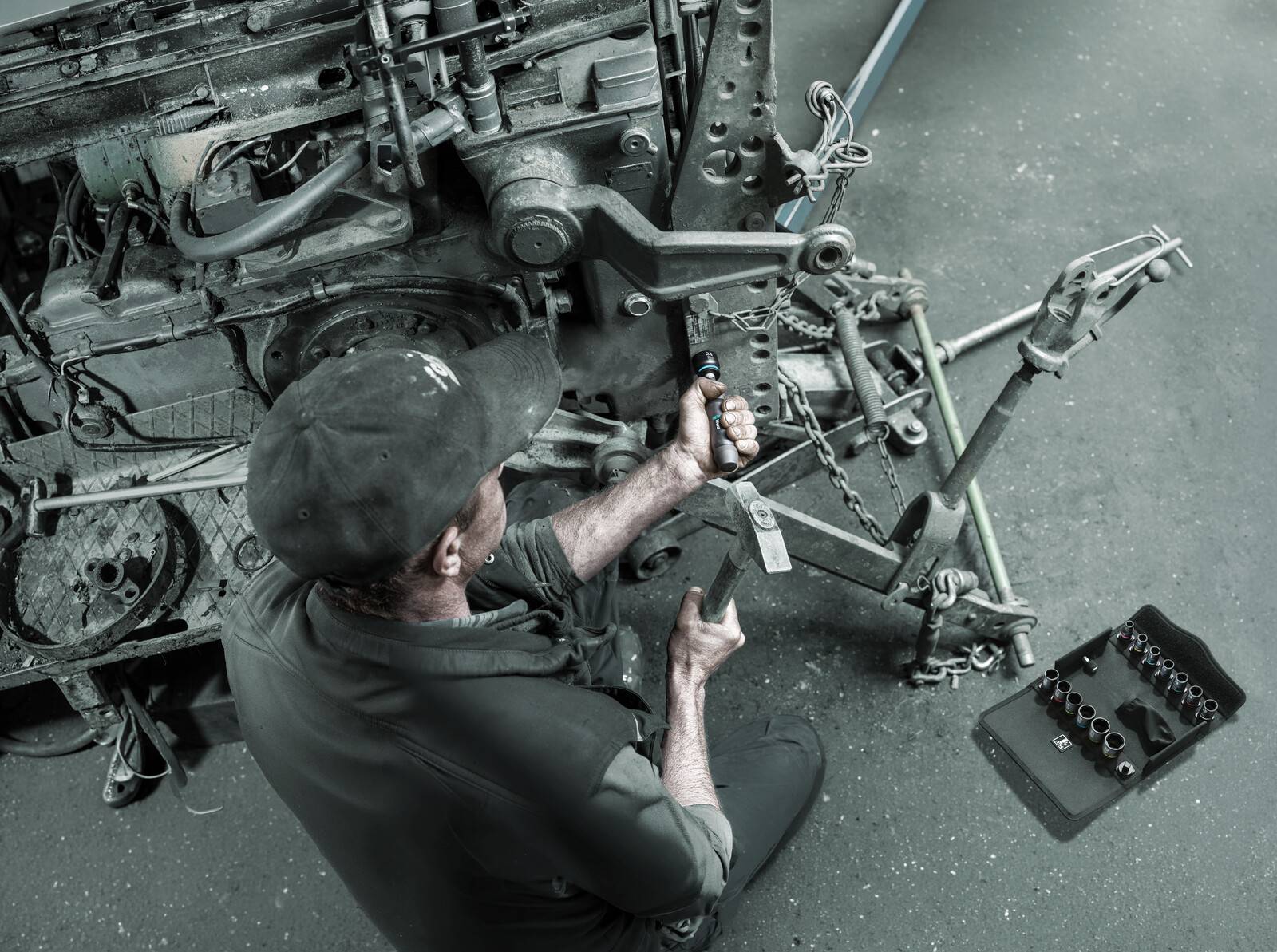 Mechaniker arbeitet an einem zerlegten Fahrzeugrahmen und nutzt ein Werkzeug in der Nähe der Vorderradaufhängung in einer Werkstatt.