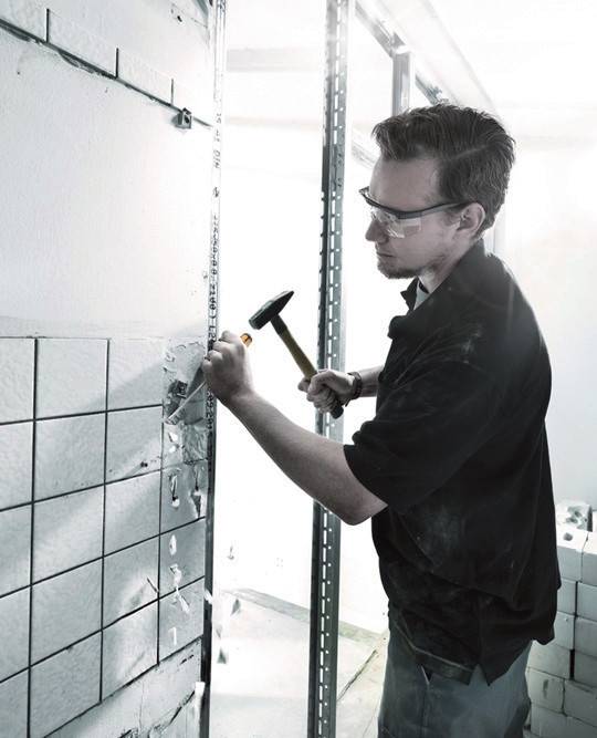 Ein Mann trägt eine Schutzbrille und verwendet einen Hammer und einen Meißel, um Wandfliesen während einer Renovierung zu entfernen.