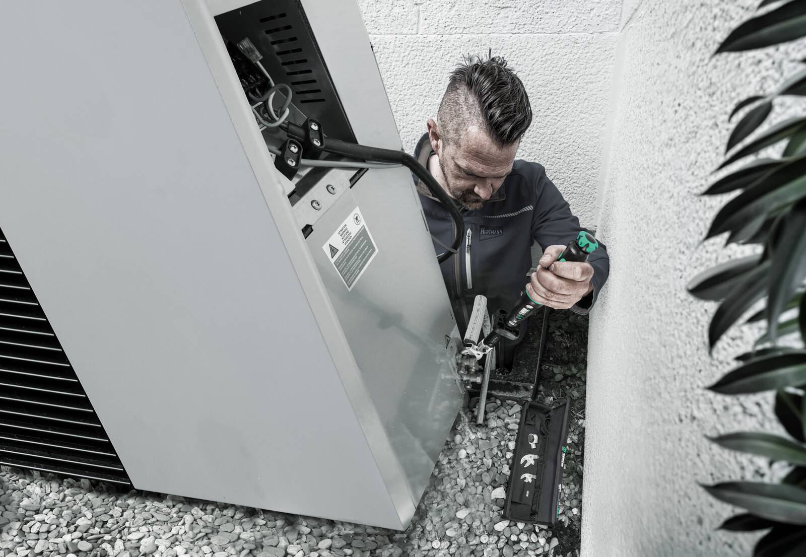 Techniker hockt neben einer Außen-Klimaanlage und repariert mit einem Elektrowerkzeug die Ausrüstung in einem engen Kieselseitenflur.