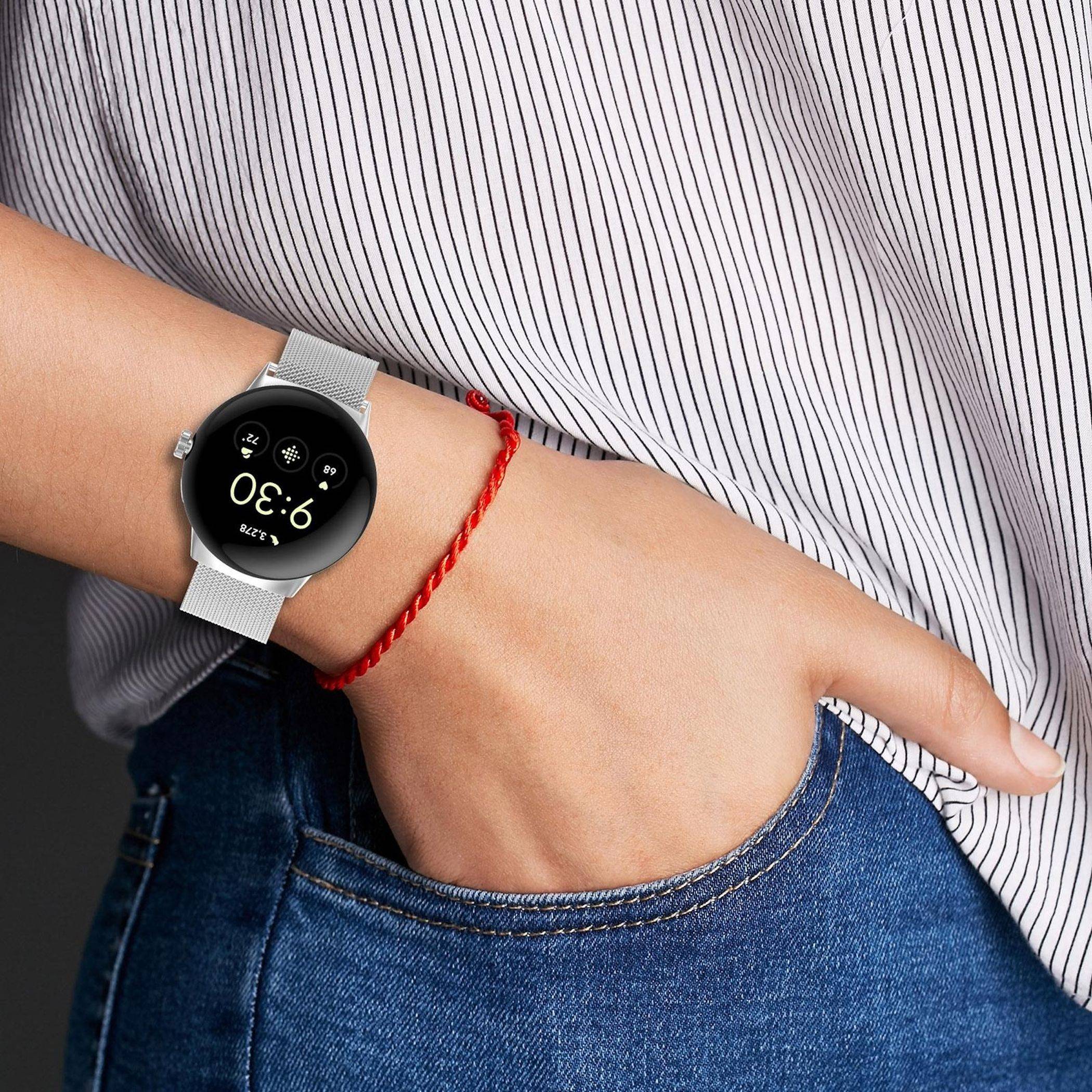 Nahaufnahme einer Person, die eine runde Smartwatch am Handgelenk trägt, mit einem roten geflochtenen Armband und einer Hand in der Jeanstasche.