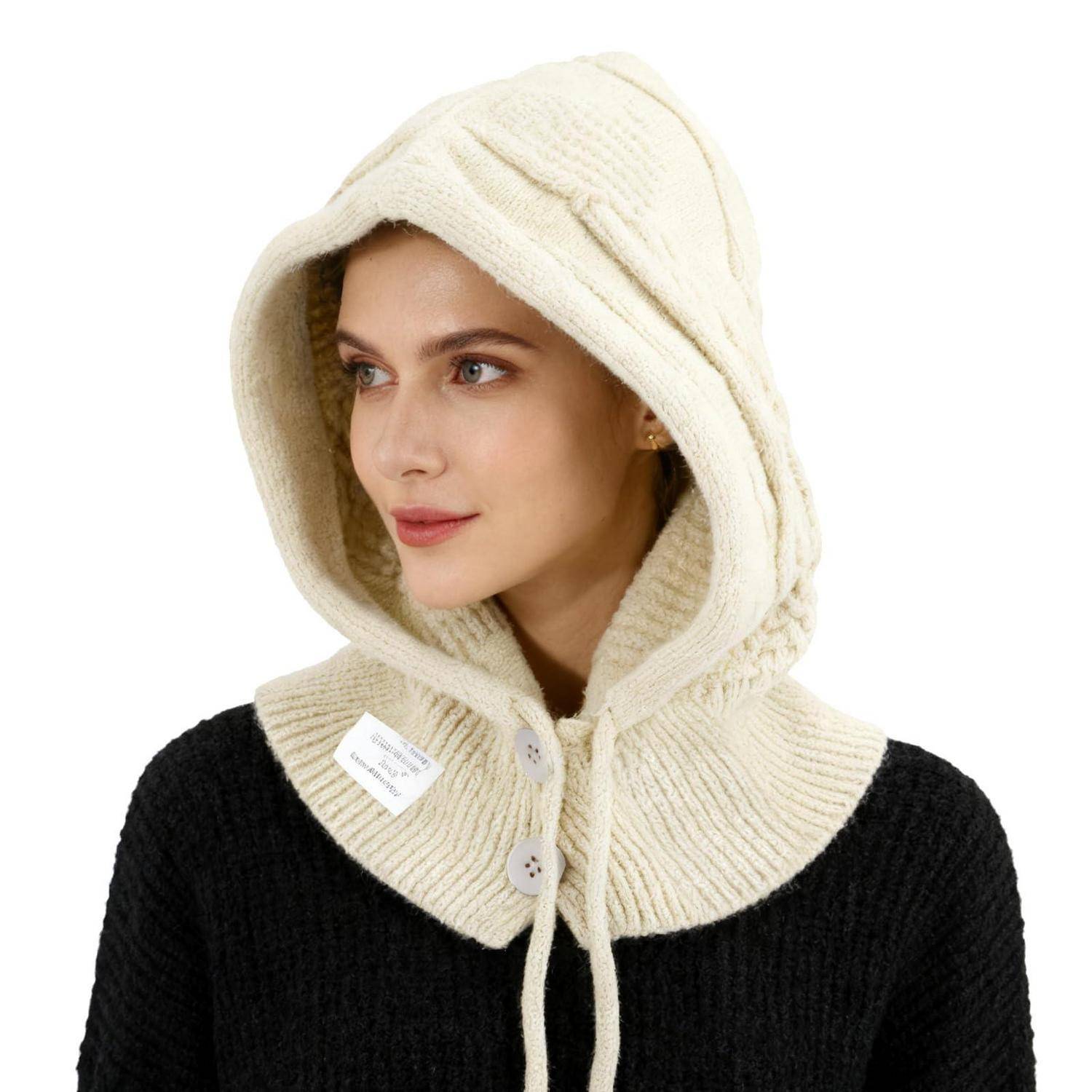 Frau, die einen cremefarbenen Strickschal mit Kapuze über einem schwarzen Pullover trägt, blickt seitlich vor weißem Hintergrund.