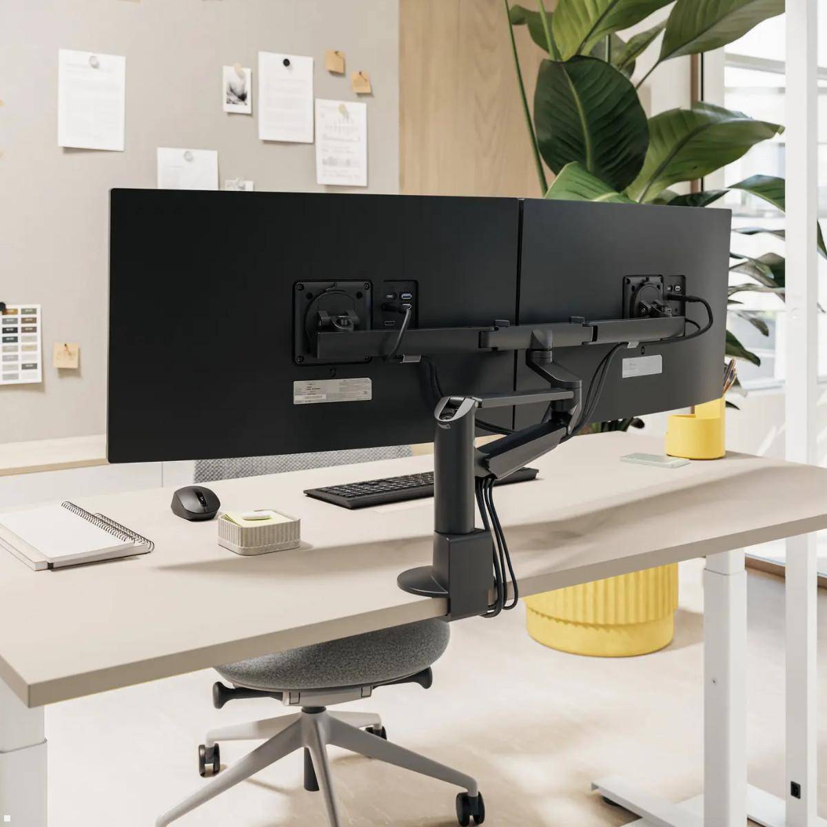 Moderner Schreibtisch mit zwei großen Monitoren auf einem verstellbaren Arm, Tastatur, Maus, Notizbuch und Stuhl in der Nähe eines Fensters und einer Pflanze.