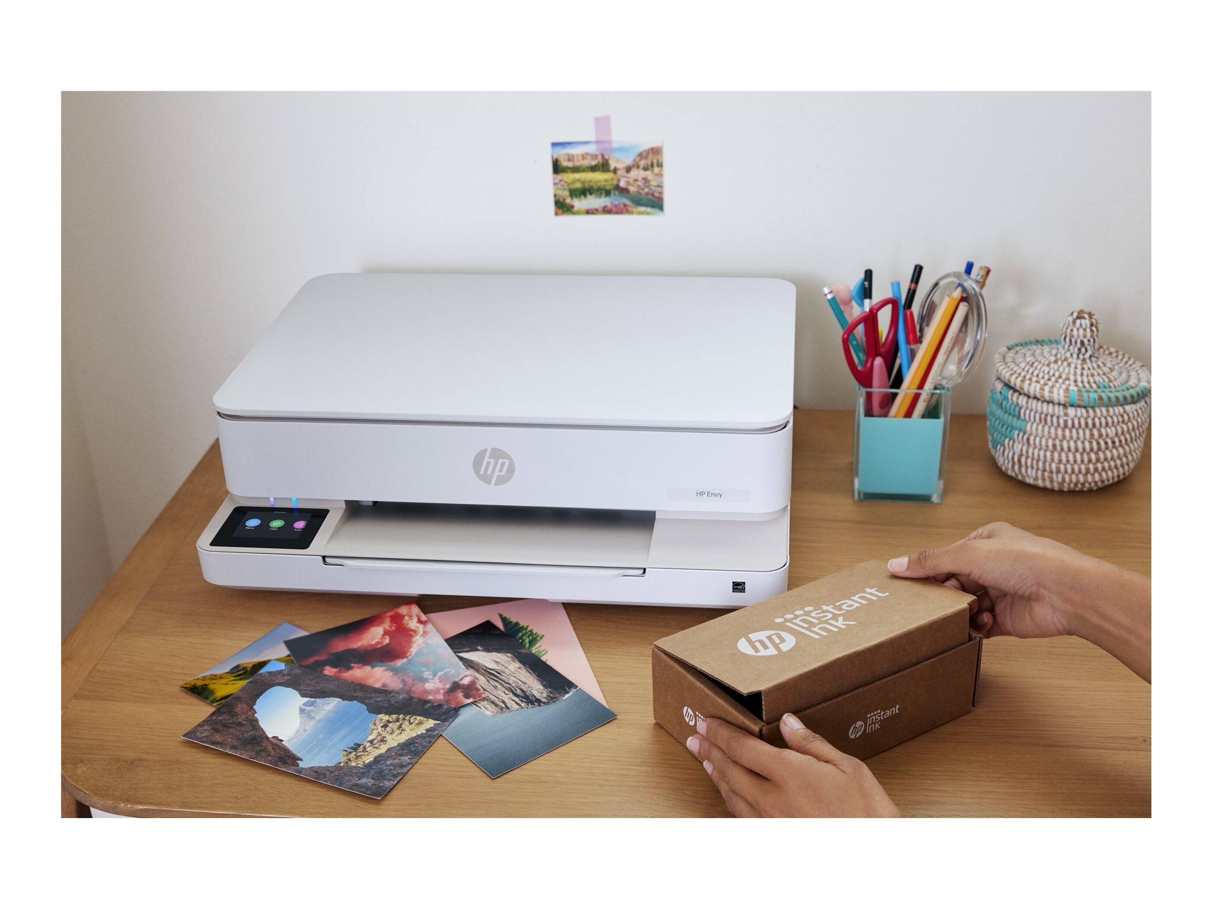 Hände öffnen eine HP-Druckerbox auf einem Schreibtisch neben einem weißen HP-Drucker, gedruckten Fotos und Schreibwarenhaltern.