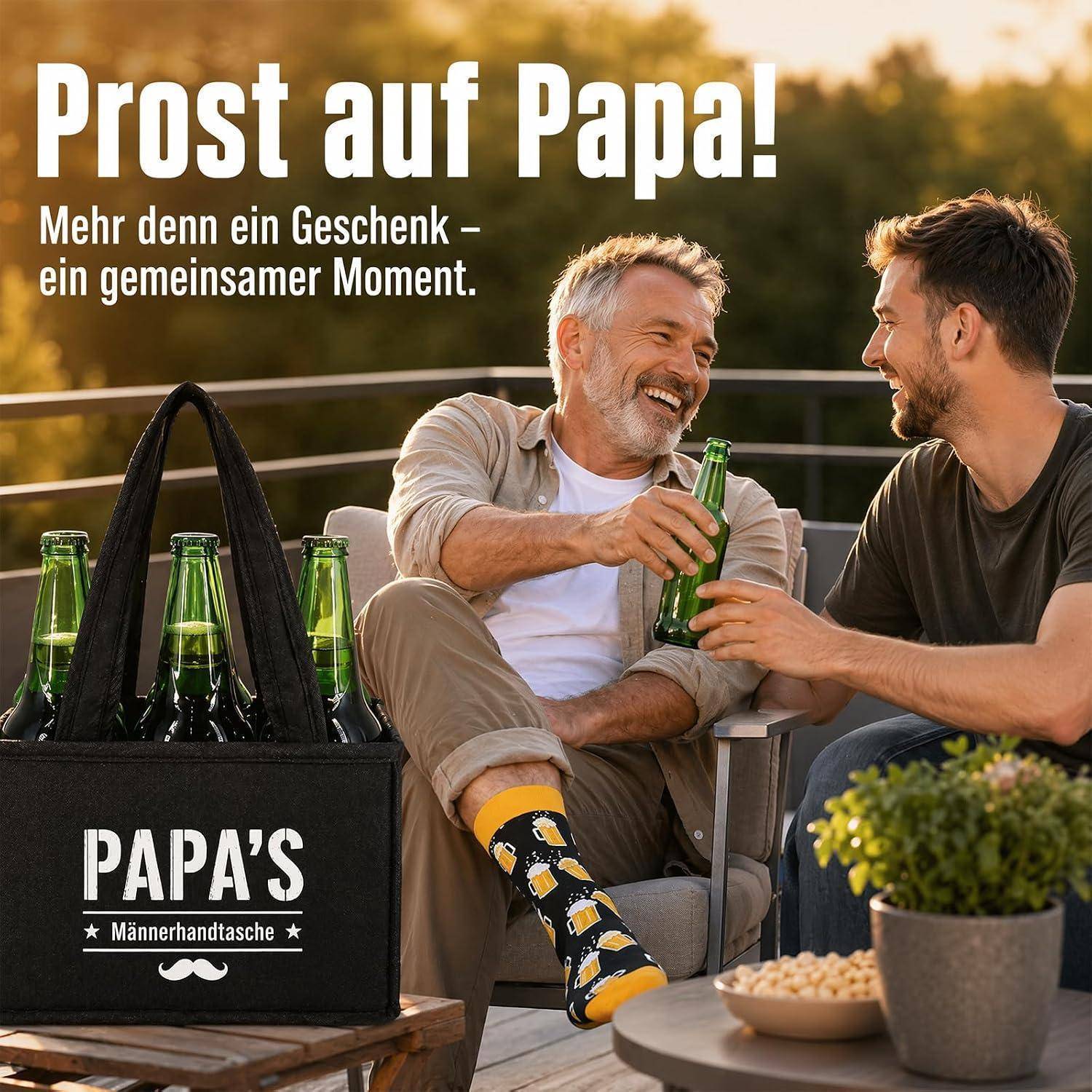 Vater und erwachsener Sohn lächeln und stoßen mit Bier im Freien an; in der Nähe eine Geschenktüte mit der Aufschrift „PAPAS Männertasche