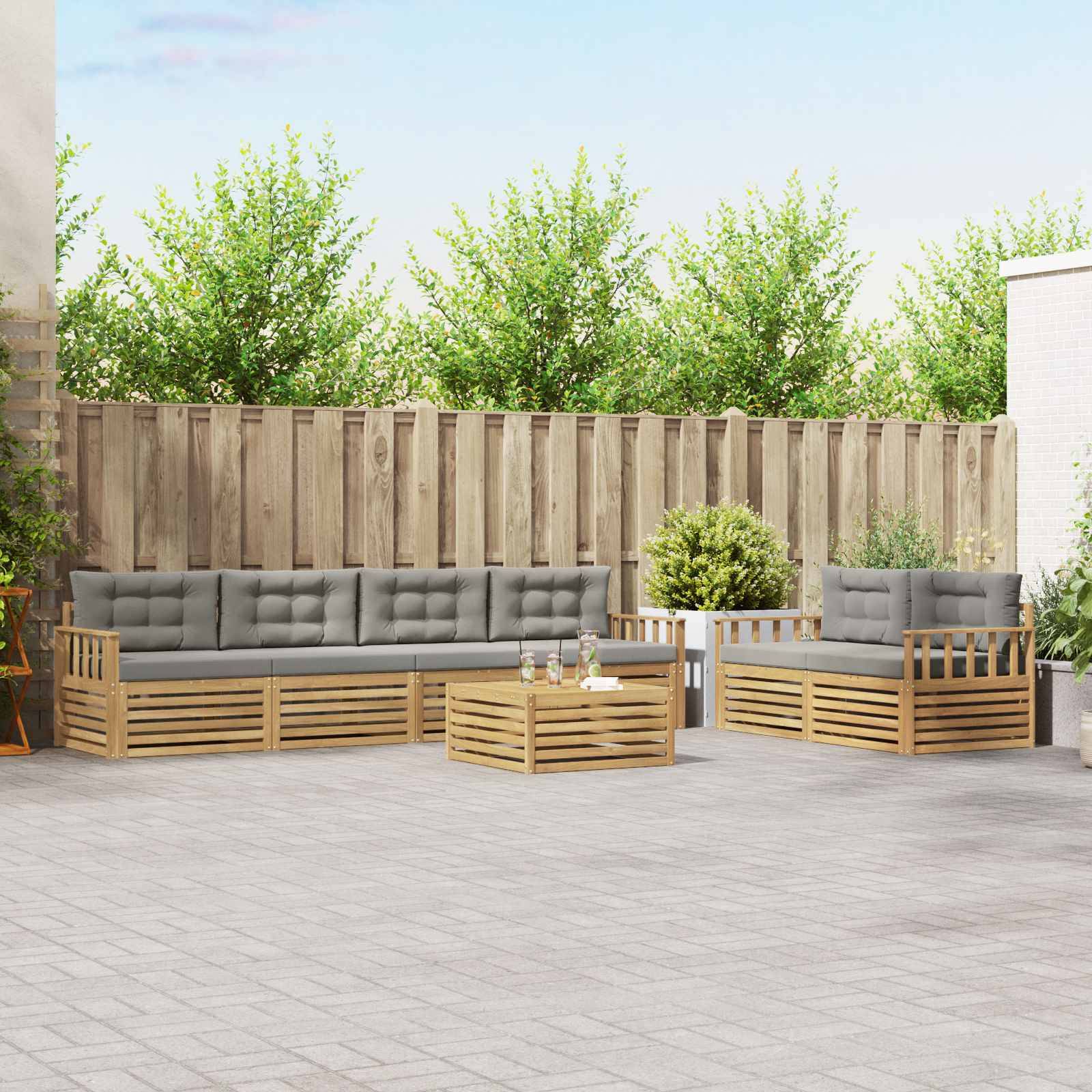 Outdoor-Sitzbereiche auf der Terrasse mit grauem Sofa und Stühlen mit Holzrahmen und Kissen um einen niedrigen Couchtisch, vor einem Holzzaun und Grünanlagen.