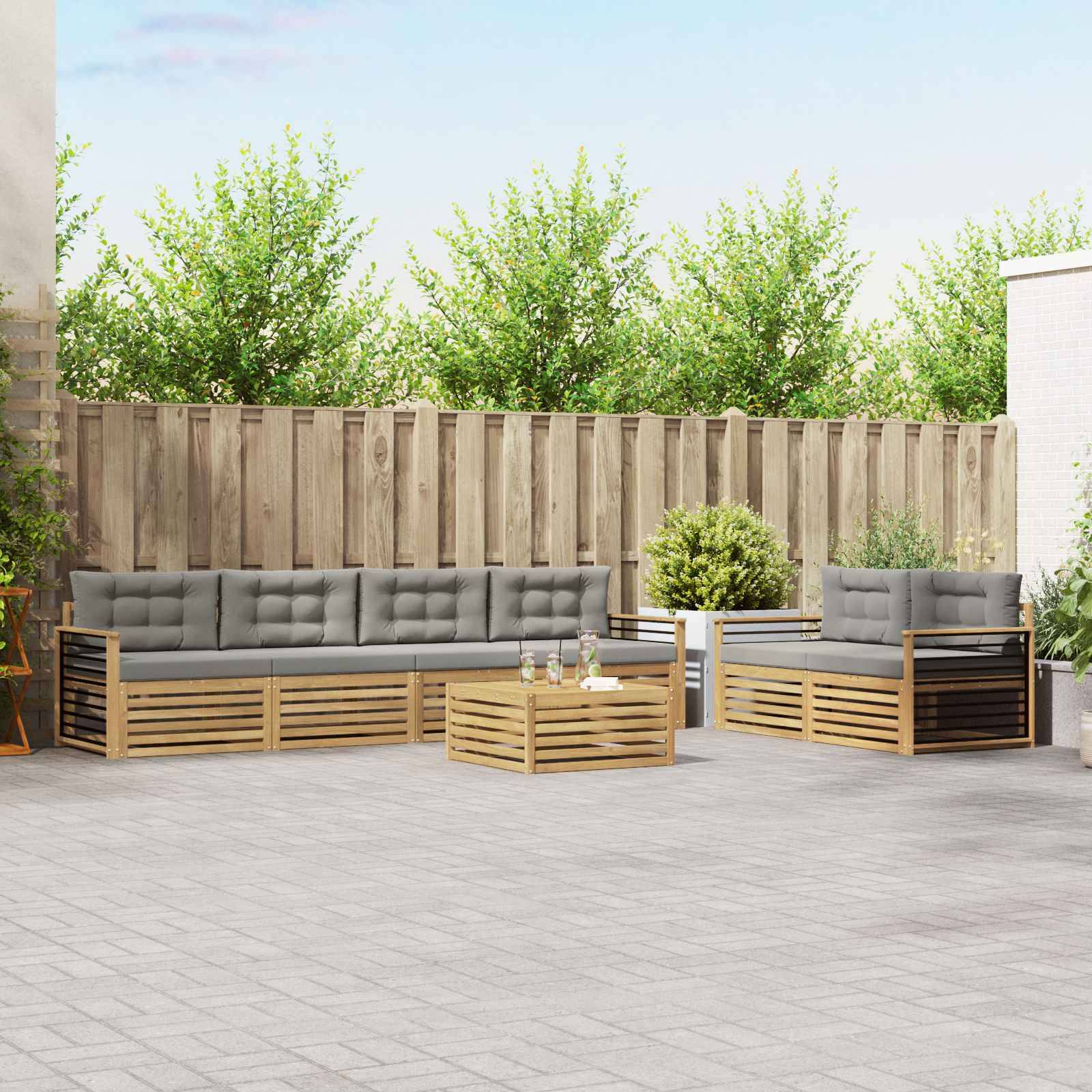 Gartenmöbel-Set für die Terrasse mit zwei grauen gepolsterten Holzsofas und einem passenden Couchtisch auf einer gepflasterten Terrasse, vor einem Holzzaun und Bäumen.