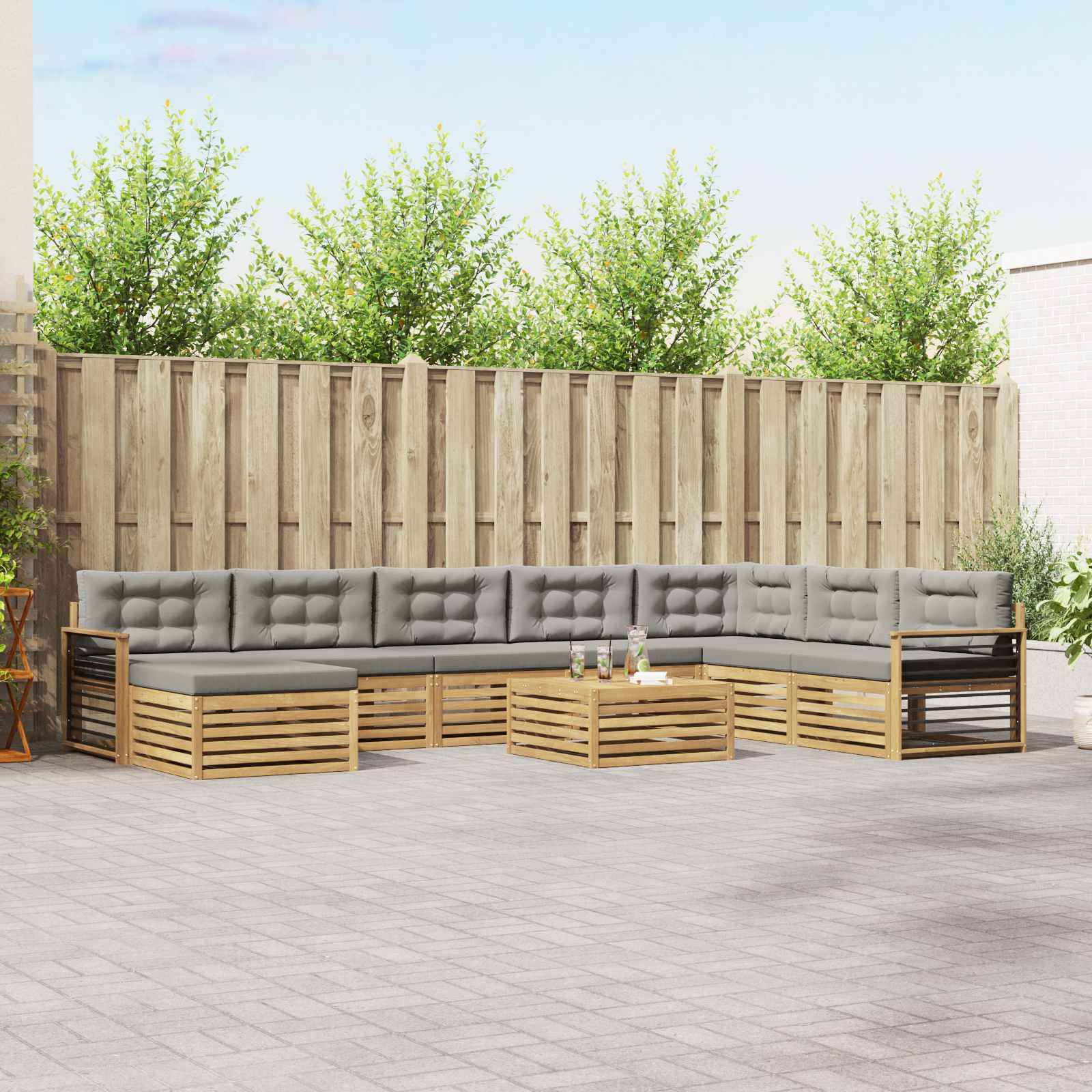 Außenterrasse mit modularer grau gepolsterter Eckgarnitur, Holzlattenrahmen und niedrigen Couchtischen vor einem hohen Holzzaun.