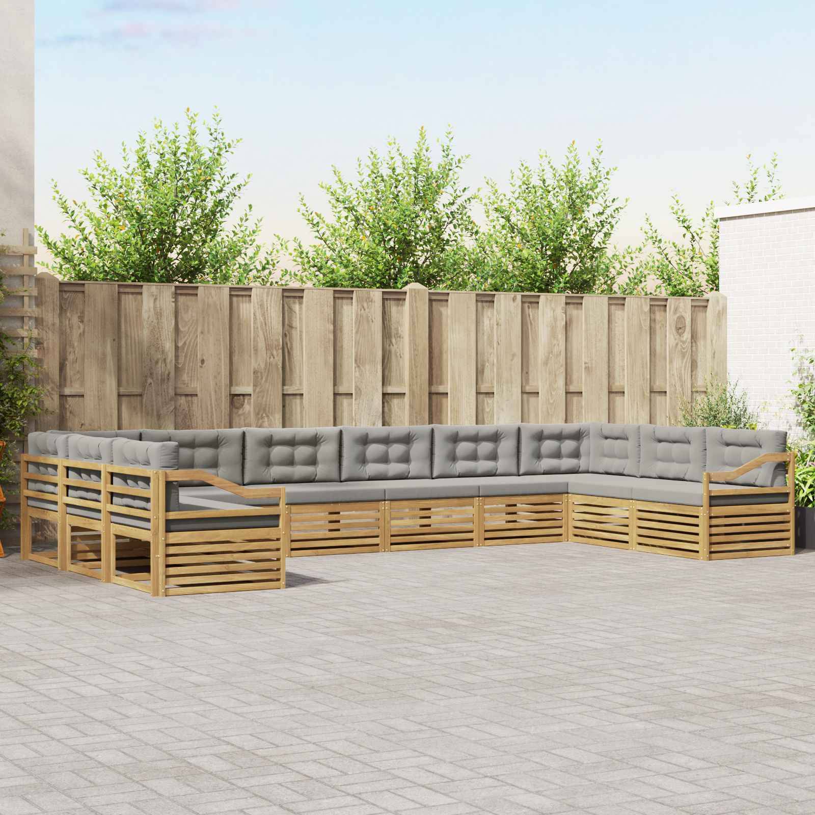 Terrasse im Freien mit einer L-förmigen Holz-Eckgarnitur und grauen Kissen vor einem Holzzaun auf einem gepflasterten Innenhof.