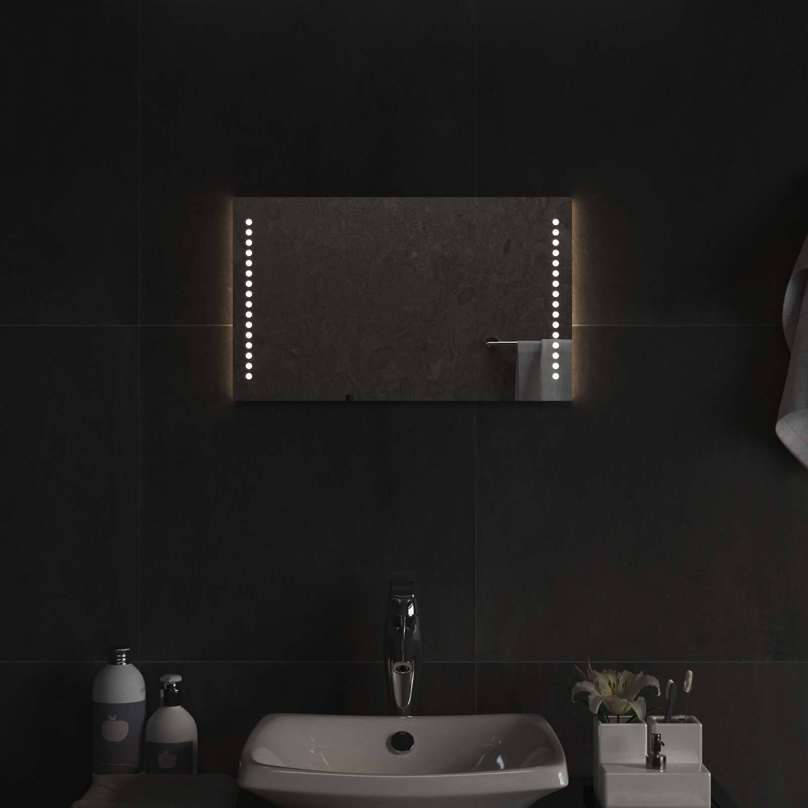 Modernes dunkles Badezimmer mit einem wandmontierten beleuchteten Spiegel über einem weißen Waschbecken und Wasserhahn, flankiert von Toilettenartikeln und kleinen Dekorationselementen.