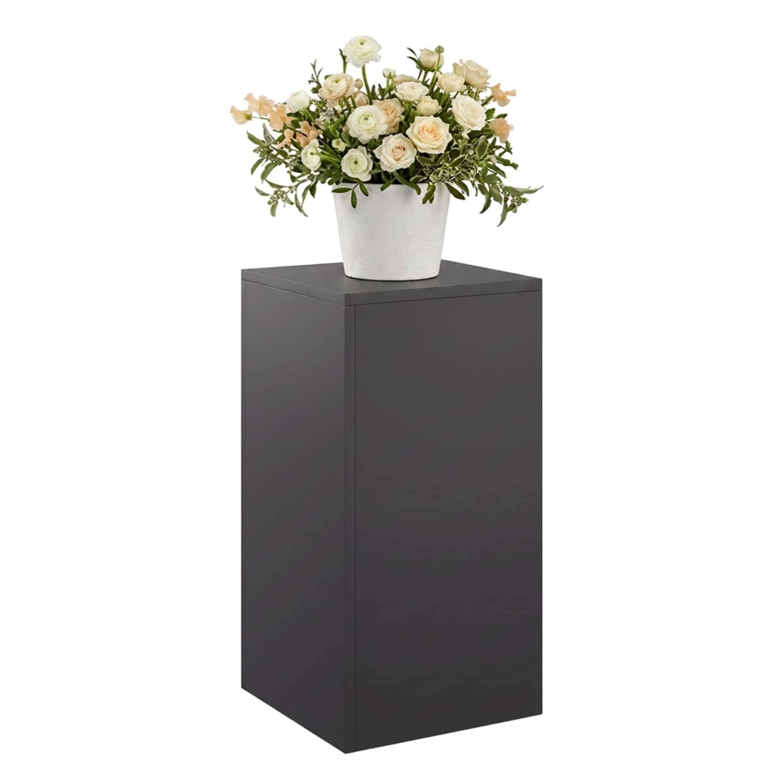 Weiße Blumengestaltung in einem weißen Topf, die auf einem hohen dunklen Sockel vor einem einfachen Hintergrund präsentiert wird.