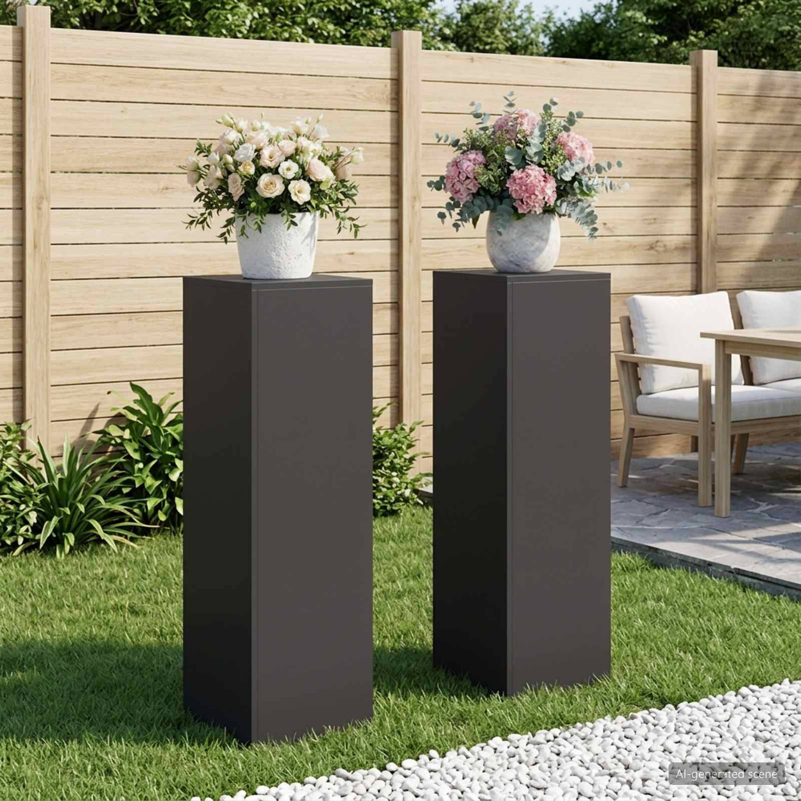 Zwei hohe dunkle Podestständer zeigen Blumenarrangements in einem gestalteten Garten neben einem Terrassensitzbereich.