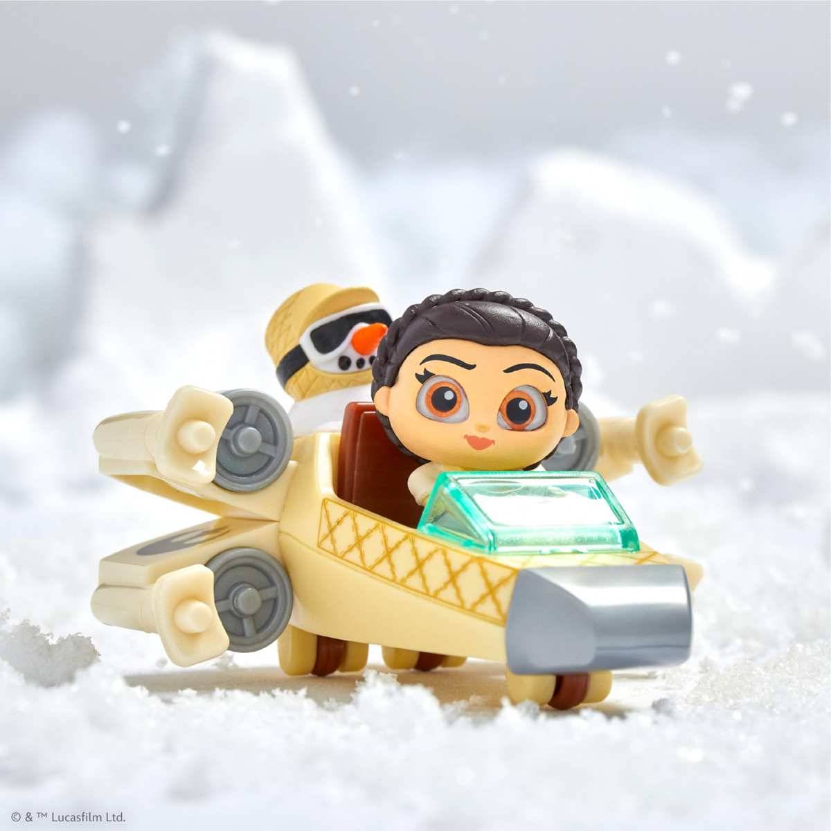 Spielzeugfiguren von Rey und BB-8, die auf einem kleinen beigefarbenen Schneegleiter auf schneebedecktem Boden fahren, mit einem sanften, eisigen Bergabhang im Hintergrund.