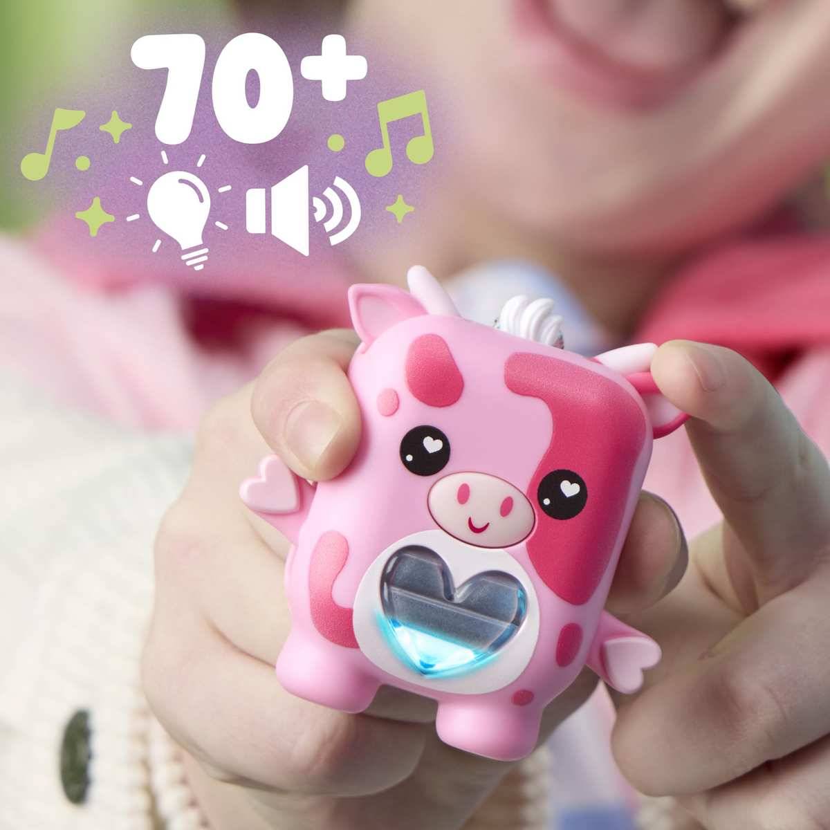 Kind hält ein rosa kuhförmiges leuchtendes Spielzeug mit einem Herzfenster; '70+' und Musik-, Licht- und Lautstärkesymbole deuten auf Geräusche und Lichter hin.