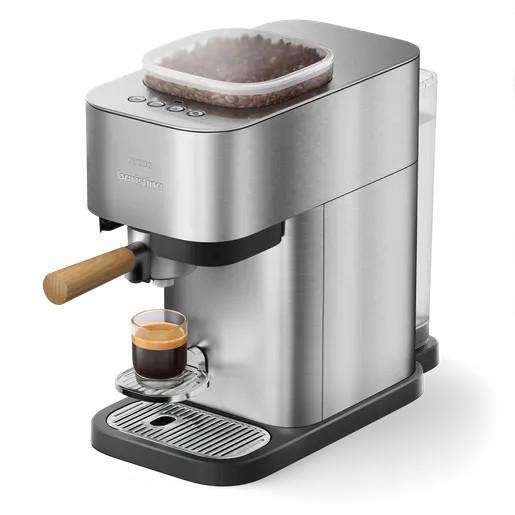 Kompakte Edelstahl-Espressomaschine, die einen Shot in eine kleine Glastasse braut, mit oberer Bohneneinfüllung und Siebträgergriff.