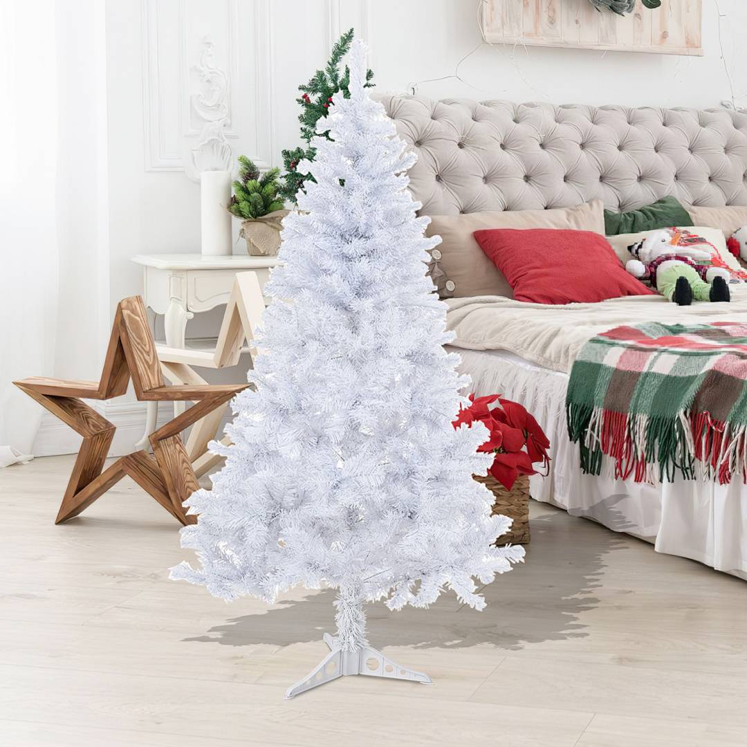 Weißer künstlicher Weihnachtsbaum in einem gemütlichen Schlafzimmer, der auf dem Boden neben einem Bett mit Weihnachtsdekoration und Kissen steht.