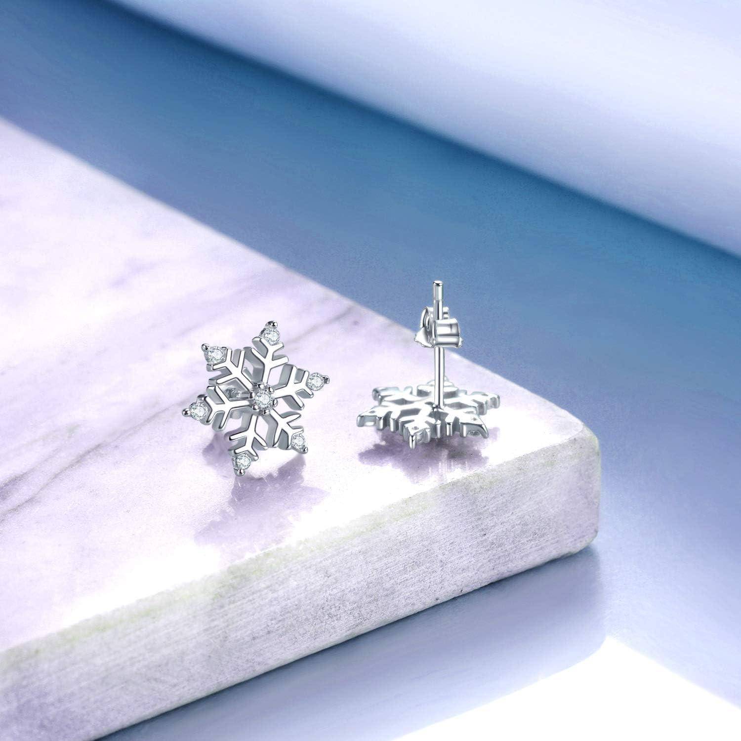 Paar silberner Schneeflocken-Stecker-Ohrringe, das auf einem hellen Marmorblock vor einem sanften blauen Hintergrund präsentiert wird.