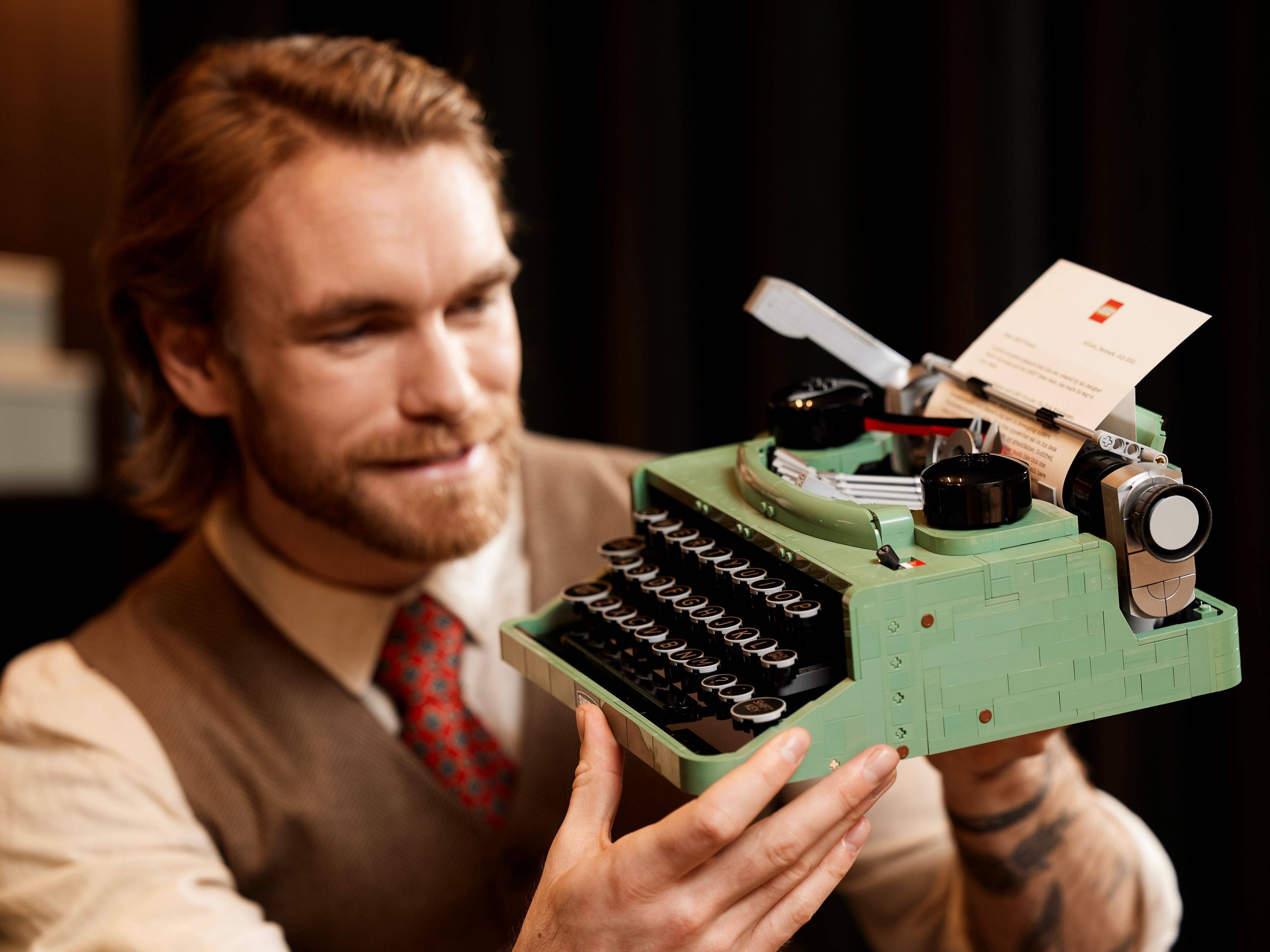 Ein Mann hält ein mintgrünes LEGO-Schreibmaschinen-Modell bewundernd in den Händen, in das ein Blatt Papier eingespannt ist.