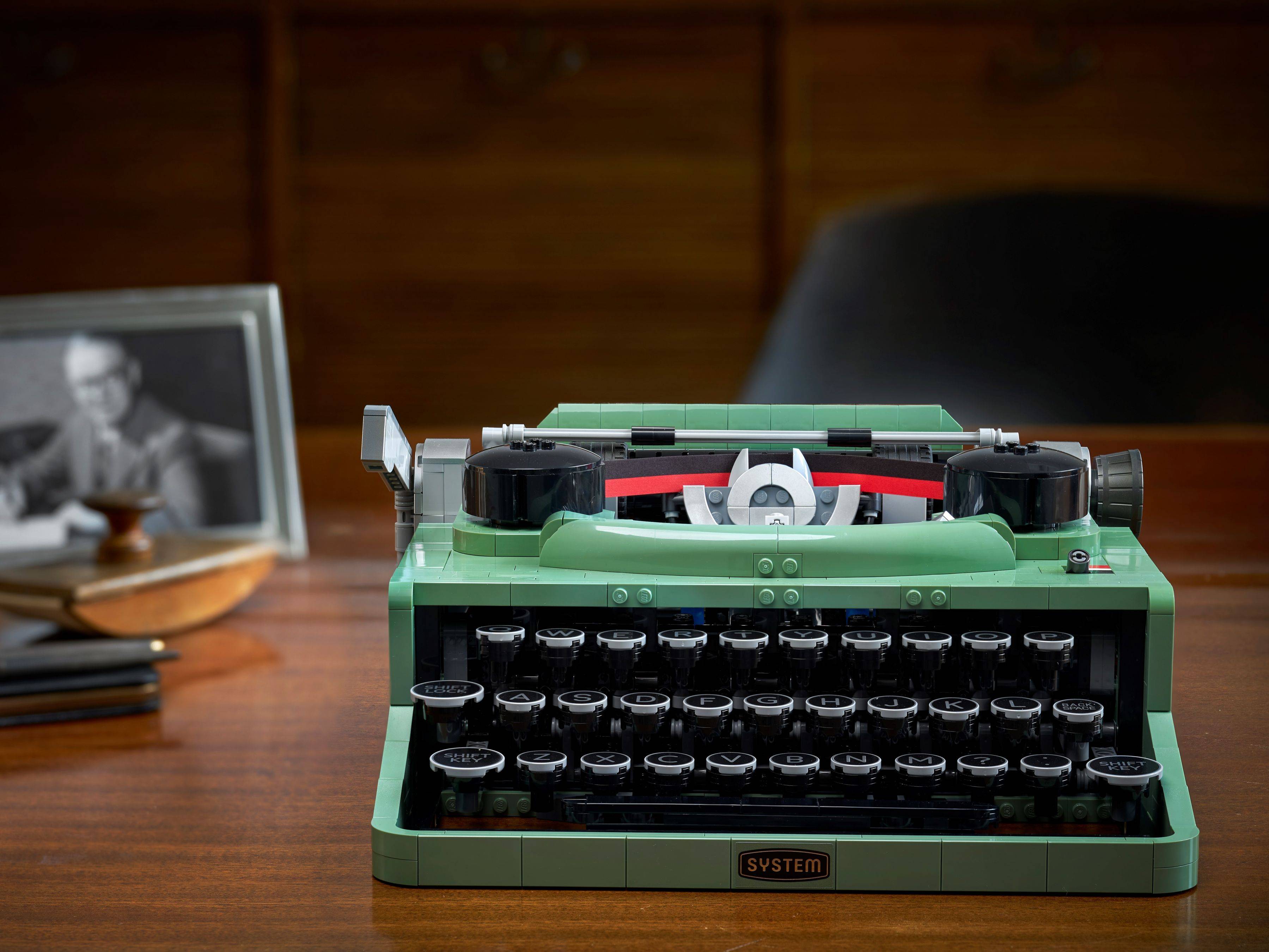 Grüne Schreibmaschine im Vintage-Stil auf einem Holzschreibtisch, mit einem unscharfen gerahmten Schwarzweißfoto und Stifthalter im Hintergrund.