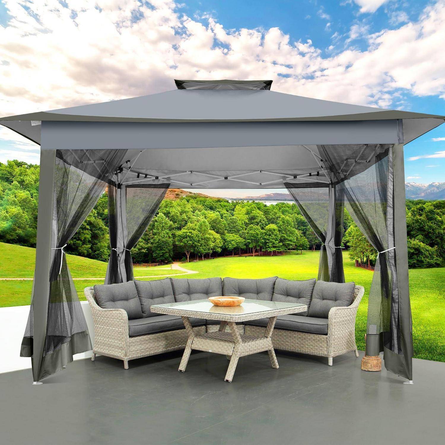 Grauer Außenpavillon mit Netzvorhängen, gepolstertes Ecksofa und Tisch auf einer Terrasse mit Blick auf einen grünen Rasen und Bäume.