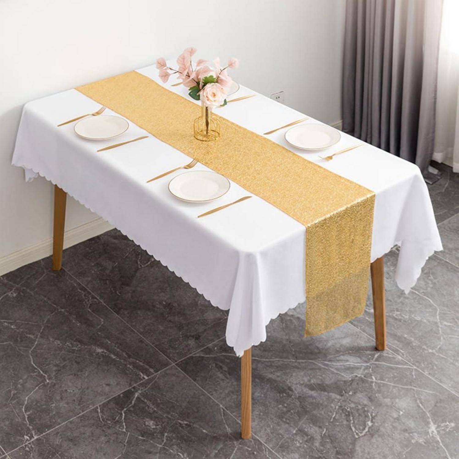 Esstisch für vier Personen mit weißer Tischdecke, goldenem Tischläufer, Tellern, Besteck und einer kleinen Blumenvase als Tischdekoration in der Mitte.