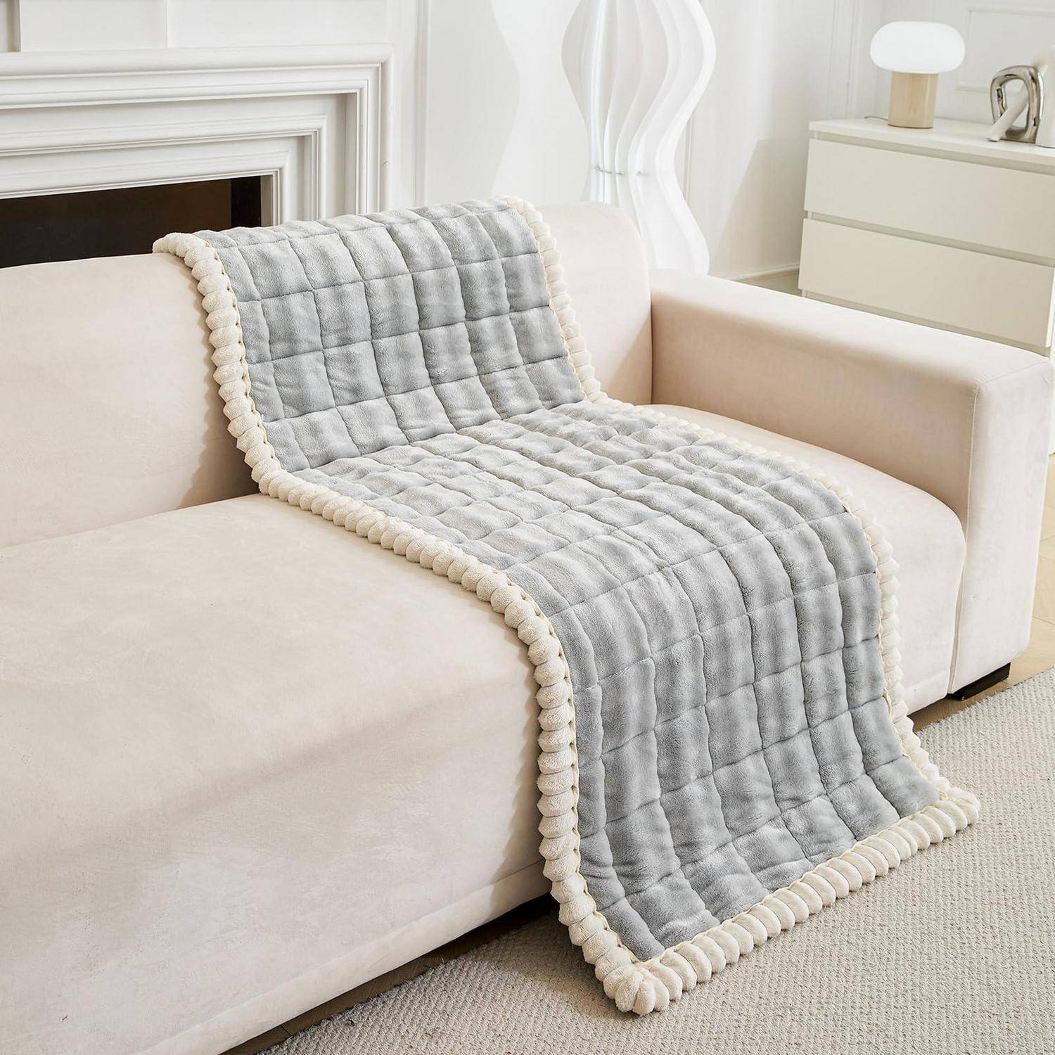 Graugedeckte Wurfdecke mit cremefarbenem Saum über einem beigen Sofa in einem hellen Wohnzimmer drapiert.