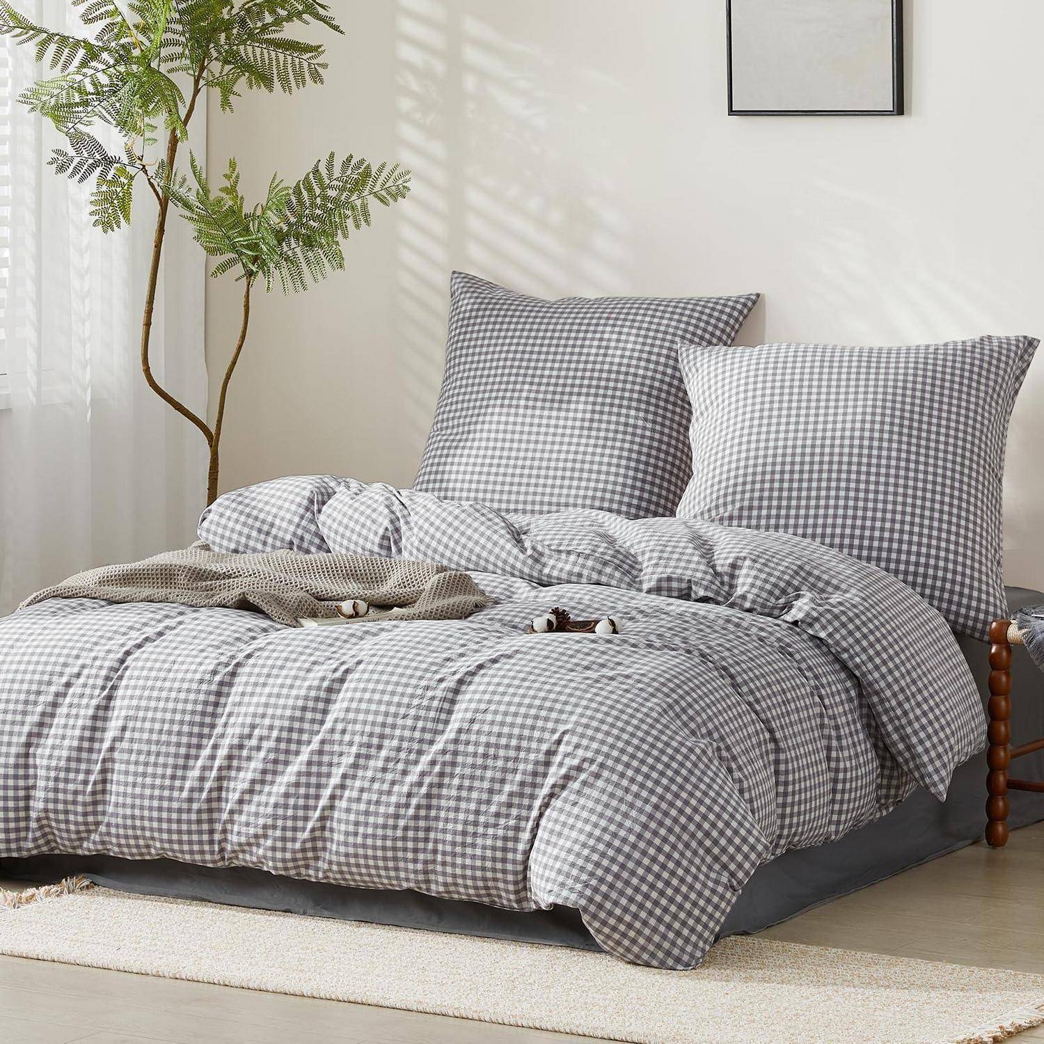 Ein ordentlich gemachtes Bett mit grauem Gingham-Bettwäsche und Kissen in einem hellen, minimalistischen Schlafzimmer mit einer Topfpflanze und leichter Wanddekoration.