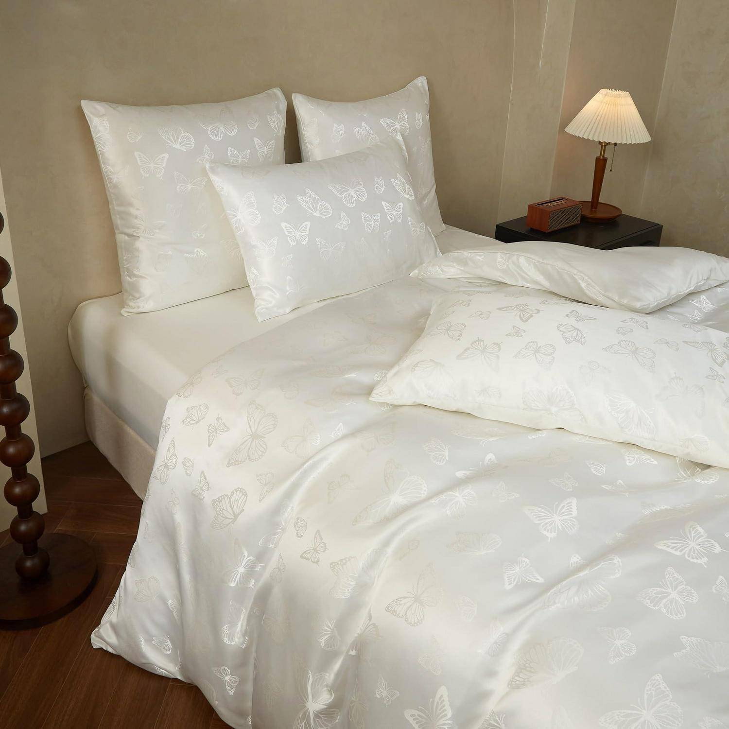 Weißes Bett mit schmetterlingsgemustertem Kopfkissen und Bettwäsche in einem sanft beleuchteten Schlafzimmer neben einer kleinen Lampe auf einem Nachttisch.