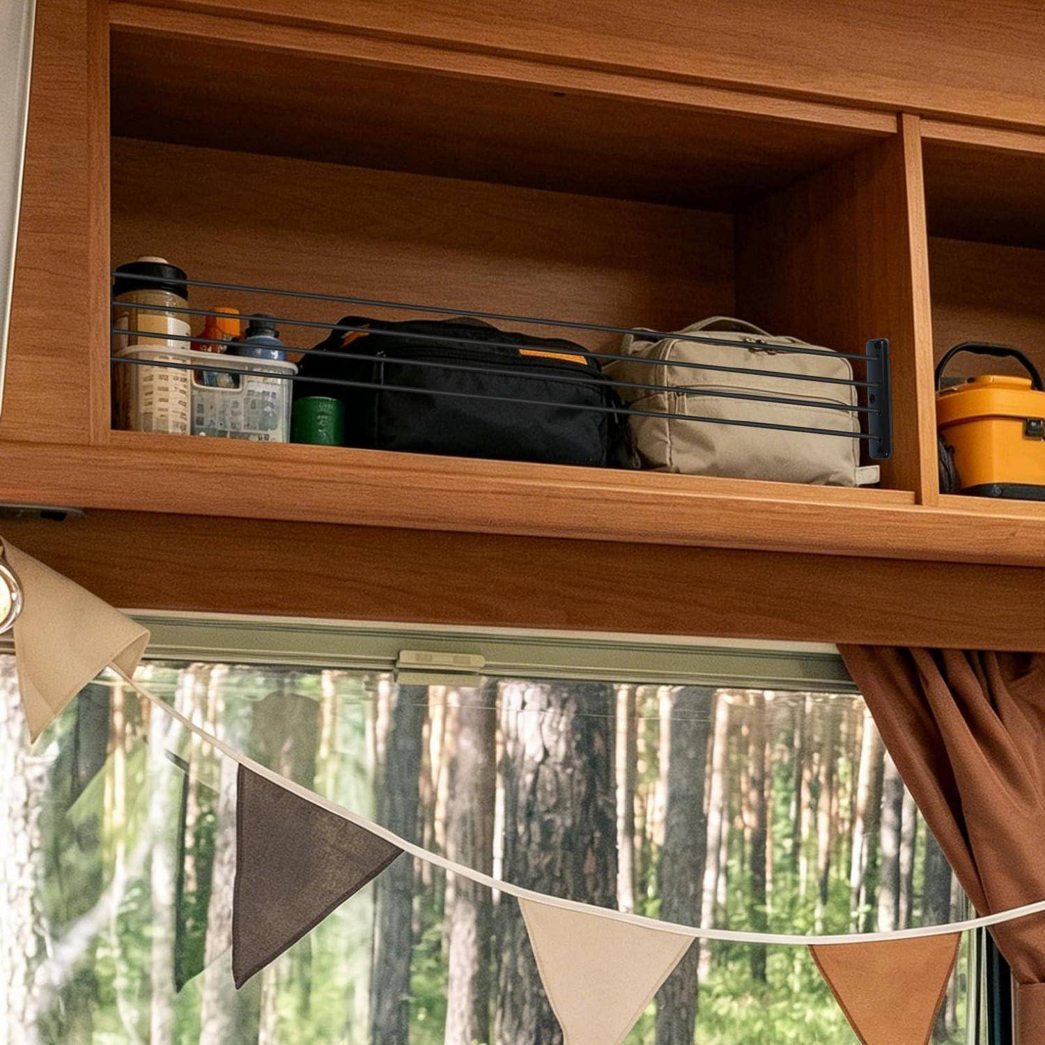Holzausstattung des Wohnmobilinneren mit Überkopf-Regalen, die Taschen und Flaschen über einem Fenster halten, das Wimpelketten, Vorhänge und einen Waldblick bietet.