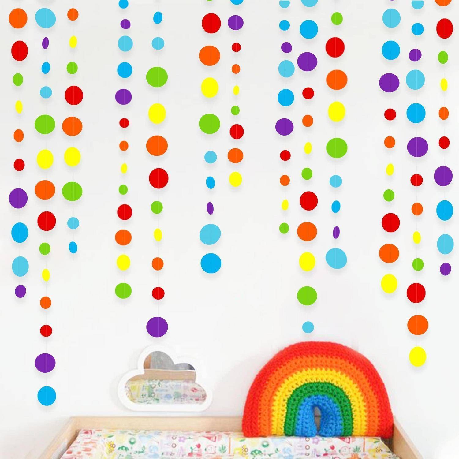 Regenbogen-Häkelgirlande auf einem Regal unter hängenden bunten Papierpunkten; ein wolkenförmiges Dekorationsstück sitzt daneben an einer weißen Wand.