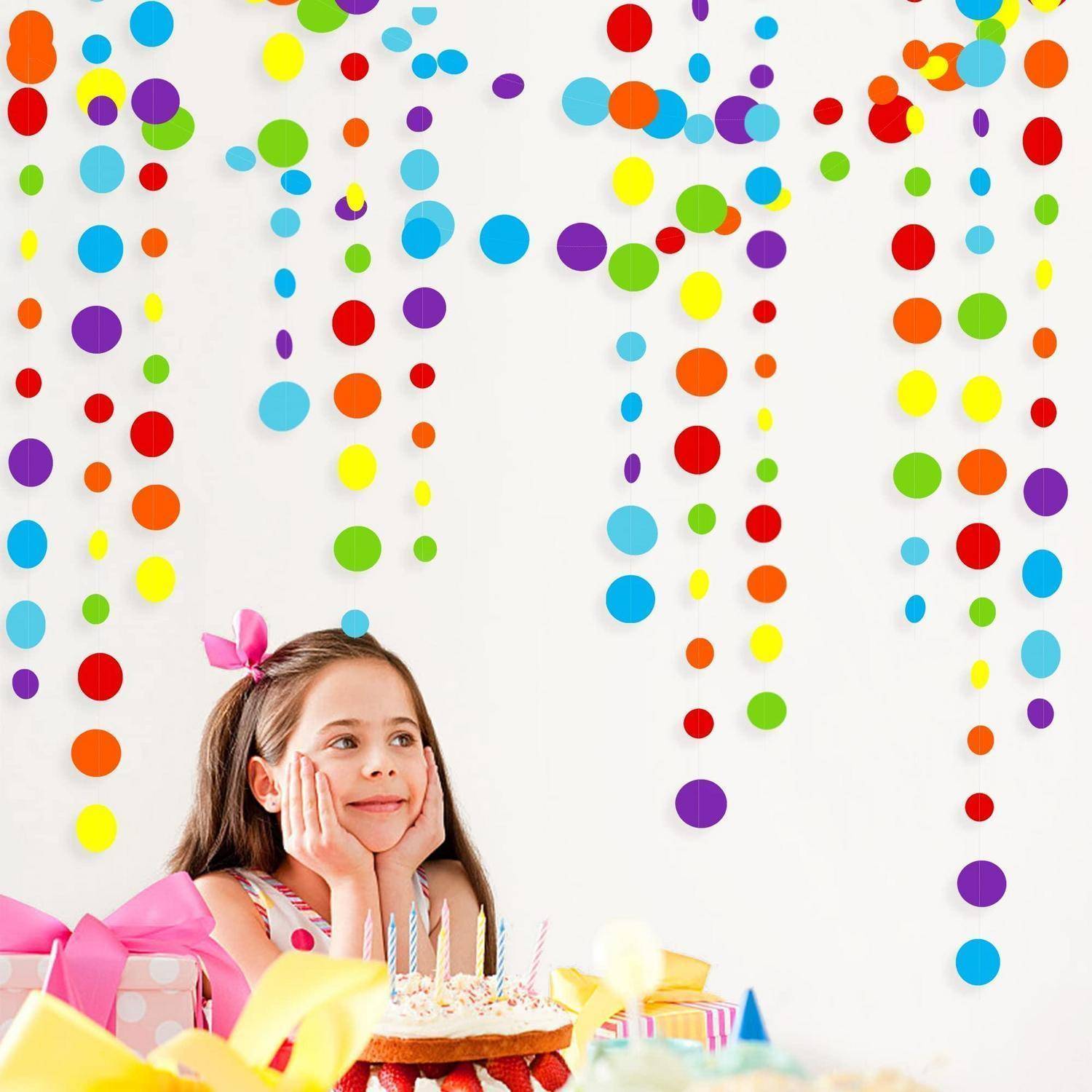Mädchen auf einer Geburtstagsfeier mit Kuchen, Kerzen und eingepackten Geschenken, das nach oben auf buntes hängendes Konfetti an einer weißen Wand blickt.
