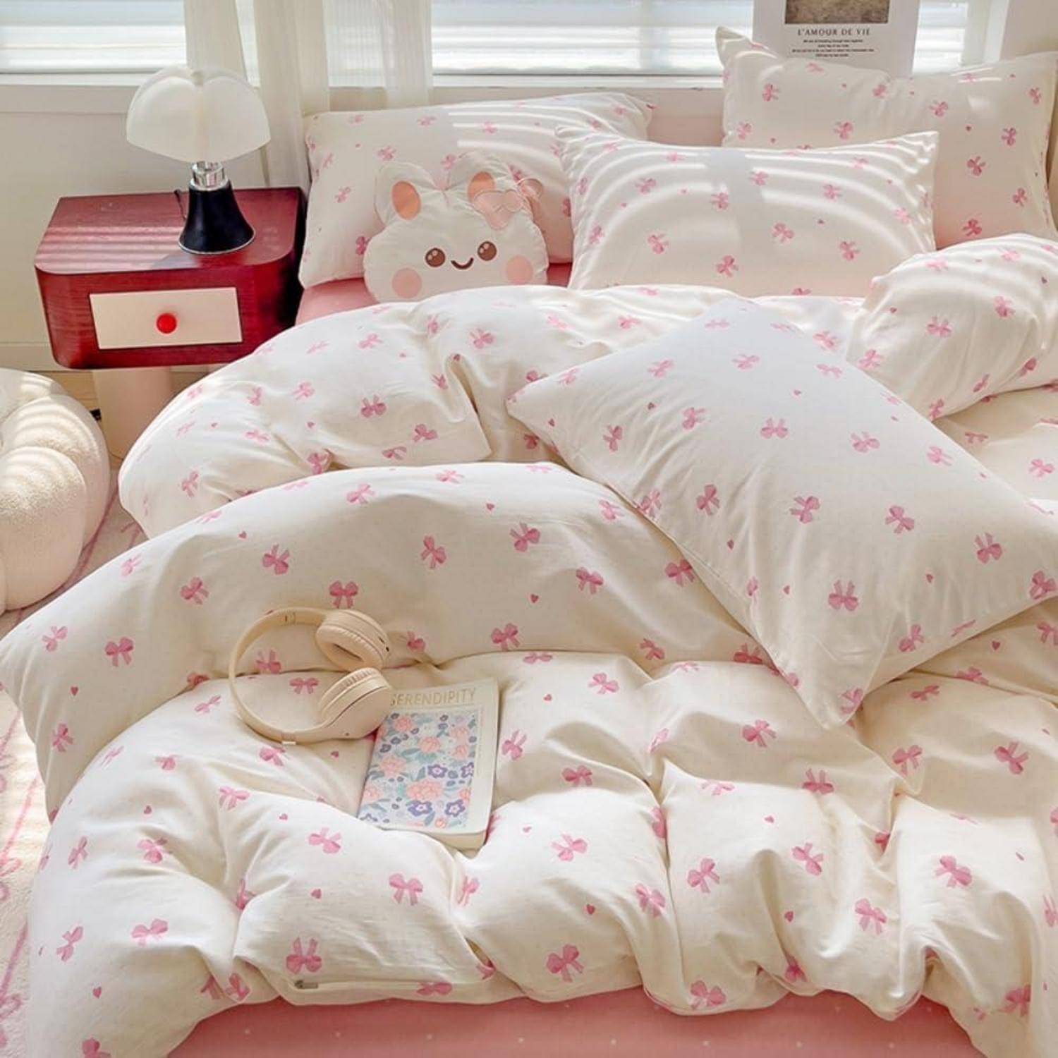 Ein ordentlich gemachtes Bett mit weiß-rosa gemusterter Bettwäsche und Kissen, auf dem ein süßes Plüsch-Gesichtskissen, Kopfhörer und ein Buch liegen.
