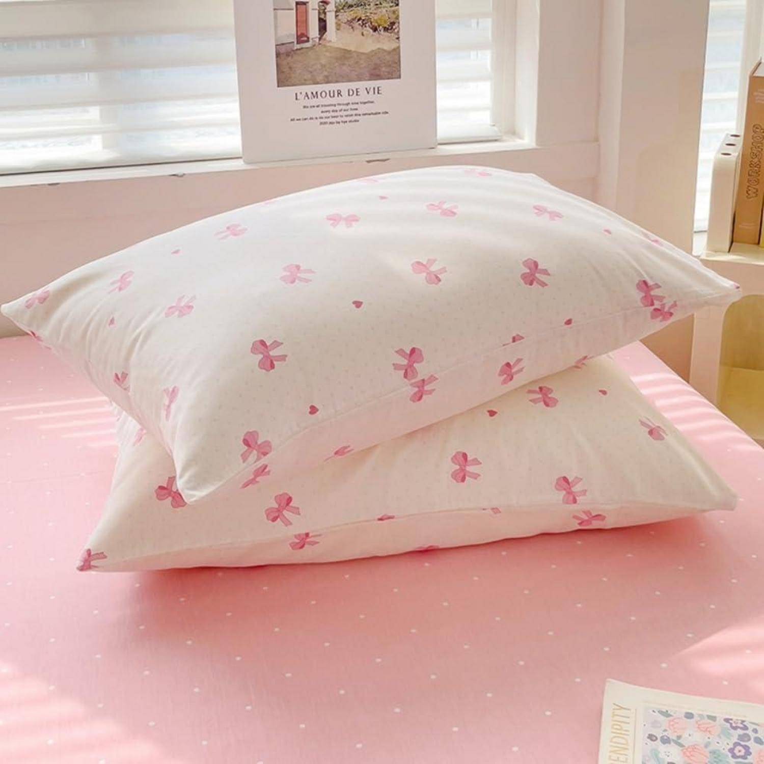 Zwei gestapelte weiße Kissen mit rosa Schmetterlingsdrucken auf einer rosa Tagesdecke in einem hellen Schlafzimmer.