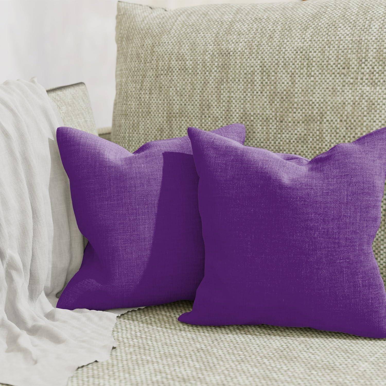 Zwei violette Wurf-Kissen auf einem beigen strukturierten Sofa neben einer weiß drapierten Decke.
