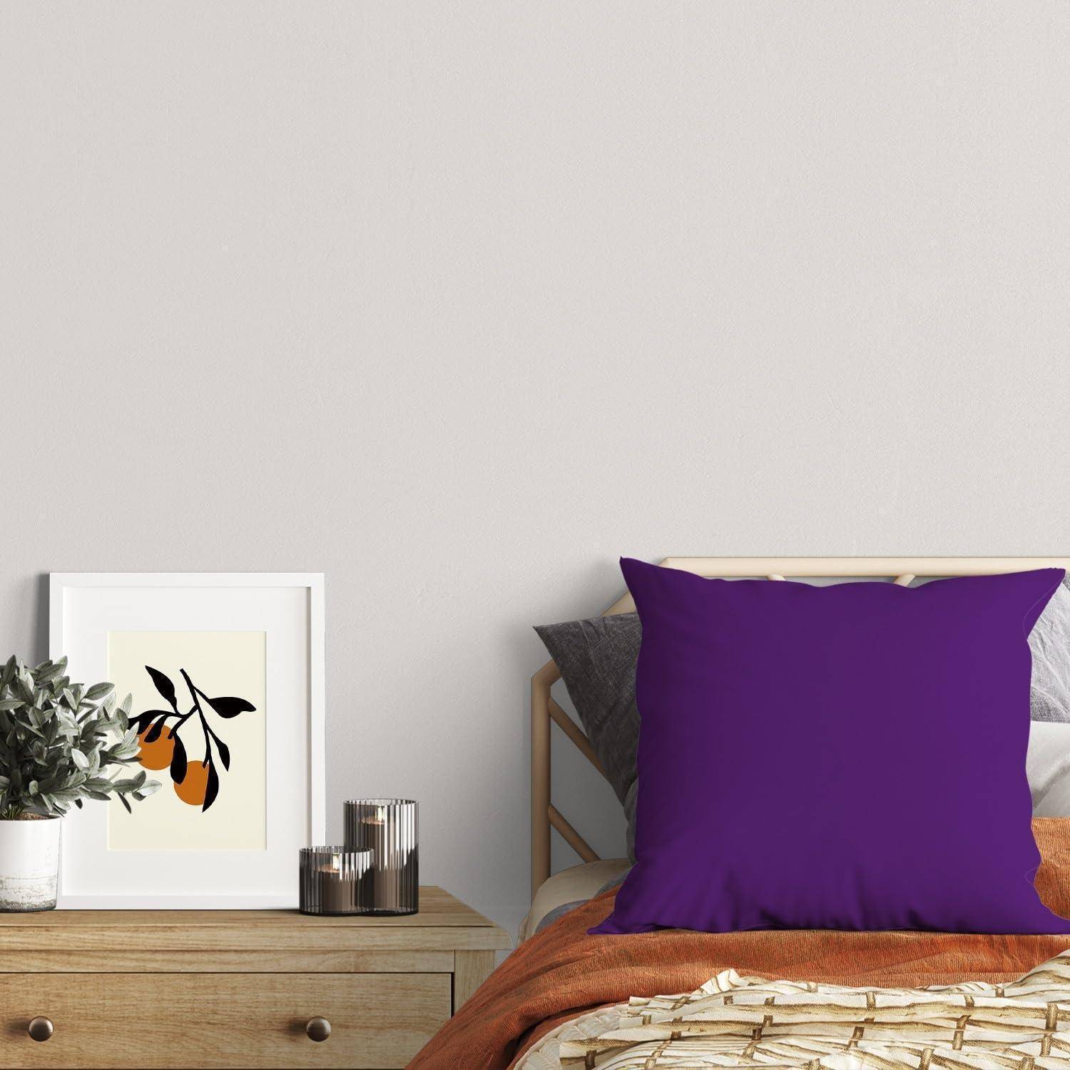 Moderne Schlafzimmerecke mit einem Bett, großem violettem Kissen, orangefarbenem Wurfstoff, gerahmter botanischer Kunst, einer kleinen Pflanze und Kerzen auf einem hölzernen Nachttisch.